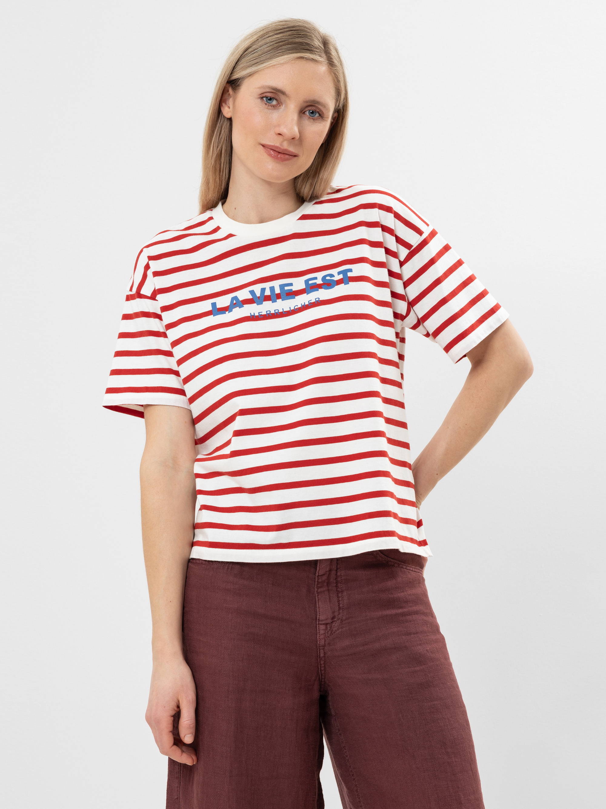 Eine Frau mit glattem blondem Haar trägt ein rot-weiß gestreiftes T-Shirt mit der blauen Aufschrift LA VIE EST. Sie trägt außerdem eine kastanienbraune Hose und steht vor einem schlichten weißen Hintergrund.
