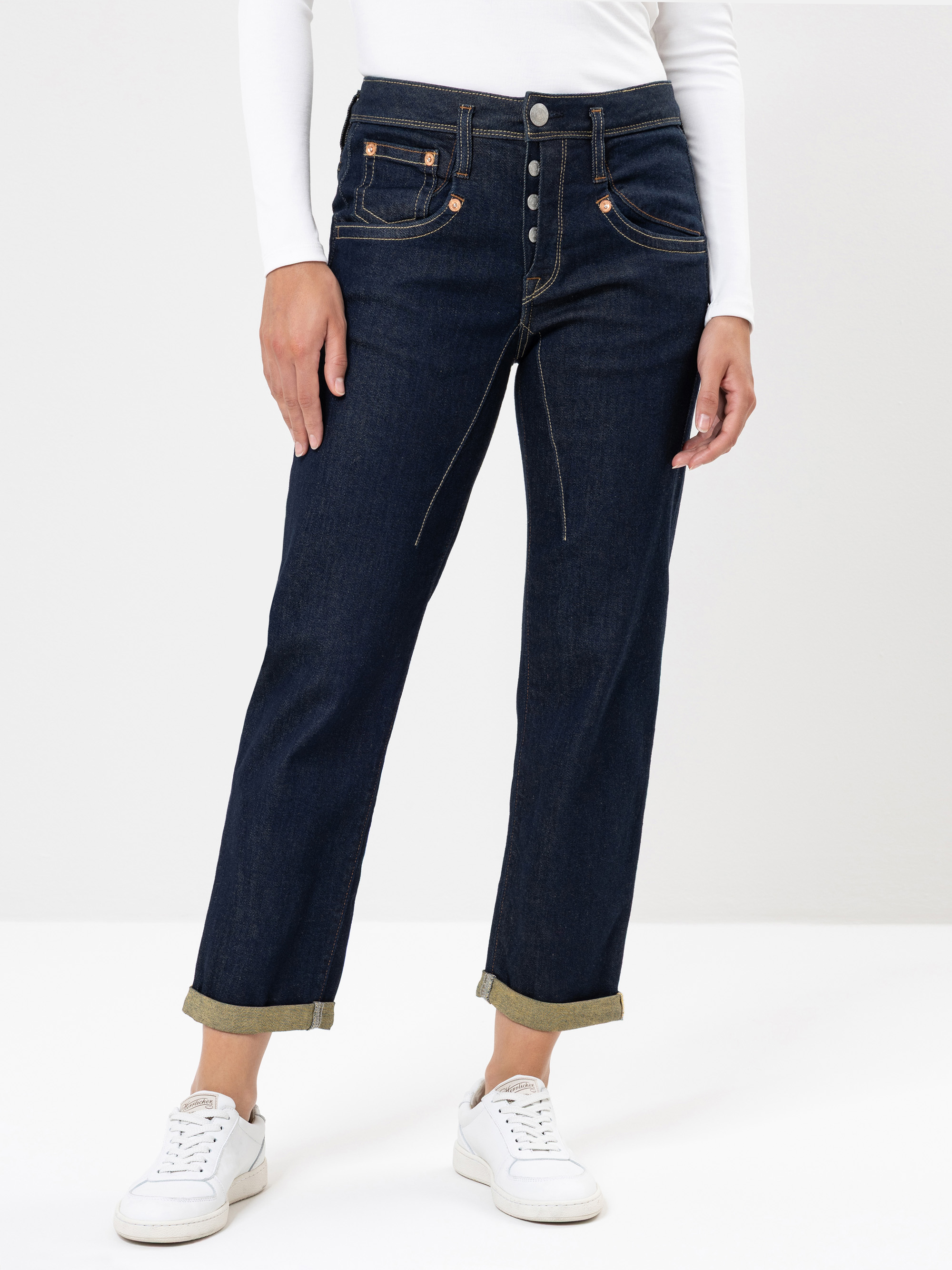 Eine Person, die eine dunkelblaue Jeans mit hoher Taille, sichtbaren Knöpfen und hochgekrempelten Manschetten trägt, dazu ein weißes langärmeliges Oberteil und weiße Turnschuhe, steht vor einem einfarbigen Hintergrund.