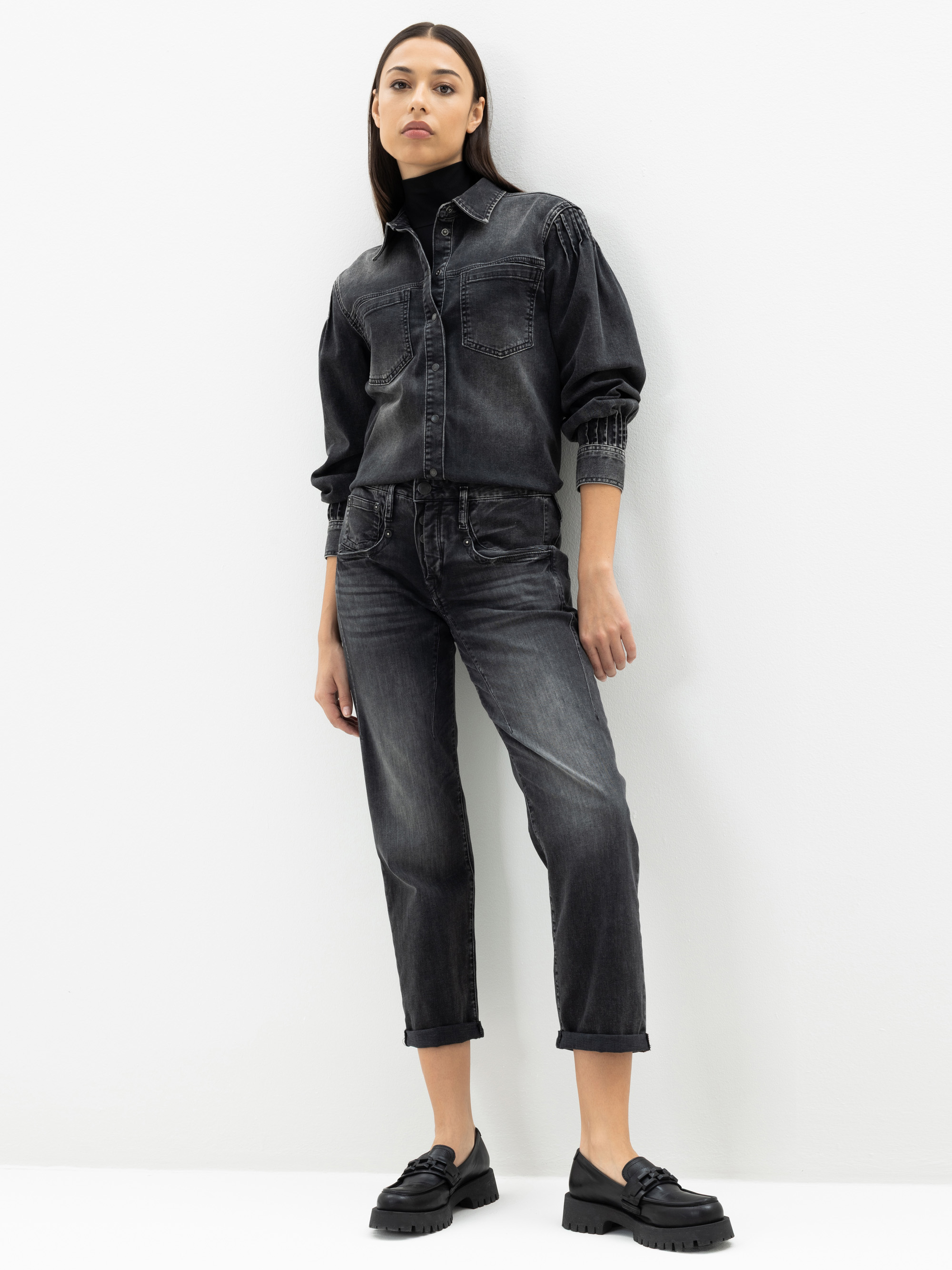 Eine Frau steht vor einer weißen Wand und trägt eine schwarze Jeansjacke und dazu passende schwarze Jeans mit schwarzen Halbschuhen. Sie hat langes, glattes Haar und einen neutralen Gesichtsausdruck.