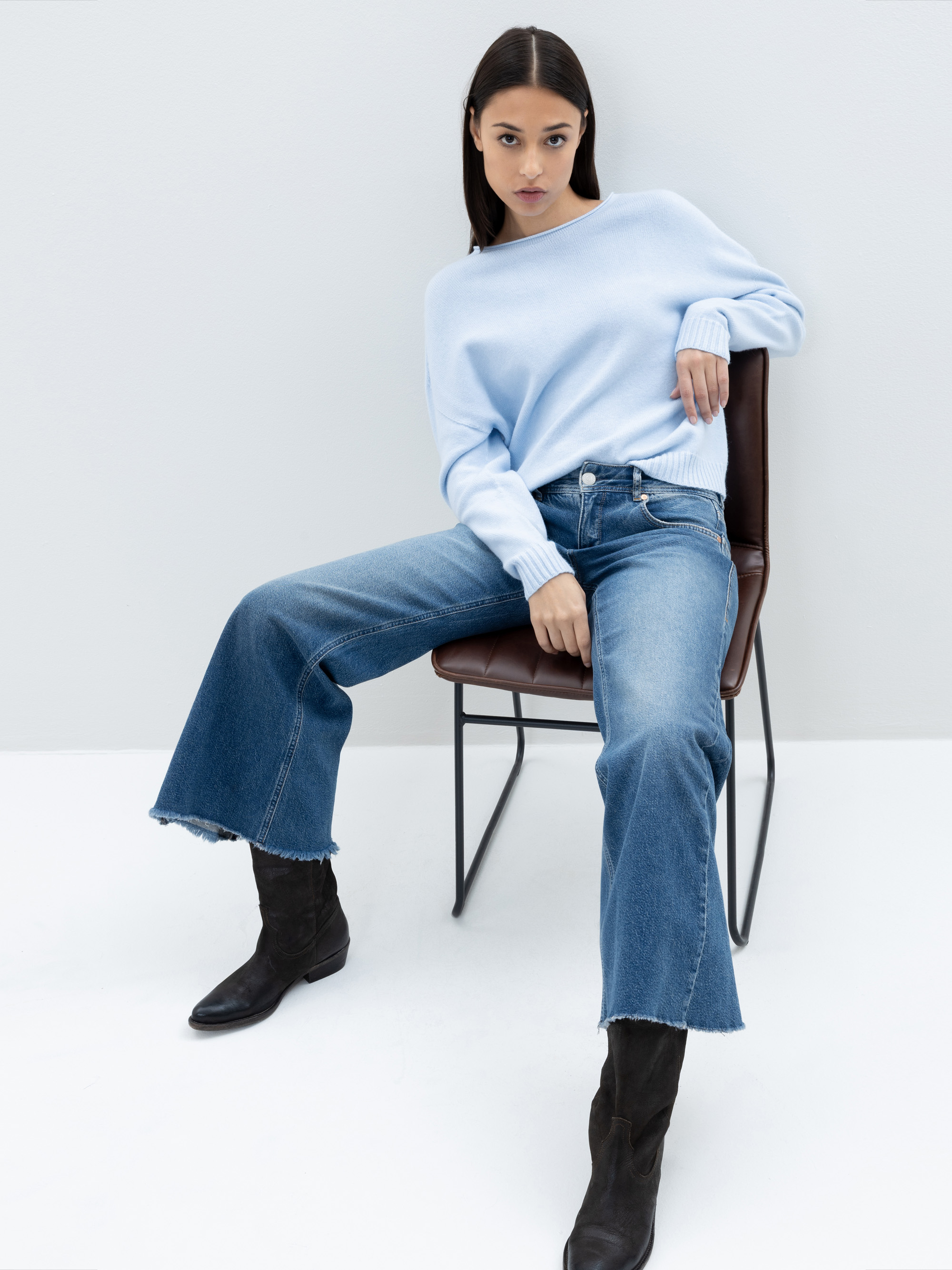 Eine Frau in einem hellblauen Pullover, weit geschnittenen Jeans und schwarzen Stiefeln sitzt auf einem Stuhl vor einem schlichten weißen Hintergrund und blickt mit selbstbewusstem Blick direkt in die Kamera.