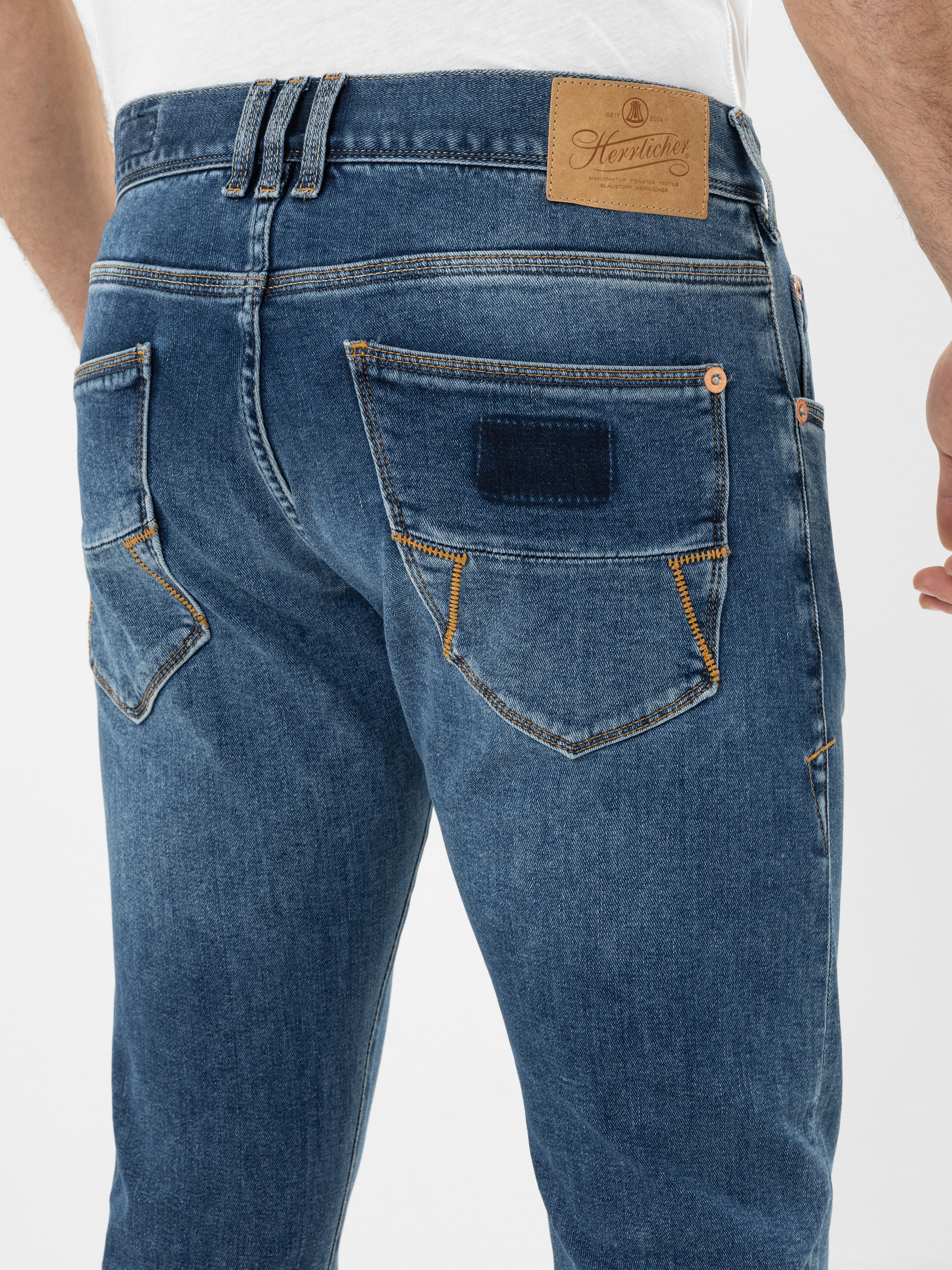 Nahaufnahme einer Person, die eine blaue Denim-Jeans mit orangefarbenen Nähten trägt. Über der rechten Gesäßtasche ist ein brauner Lederaufnäher mit der Marke Herrlicher aufgenäht. Die Person trägt ein weißes Hemd.