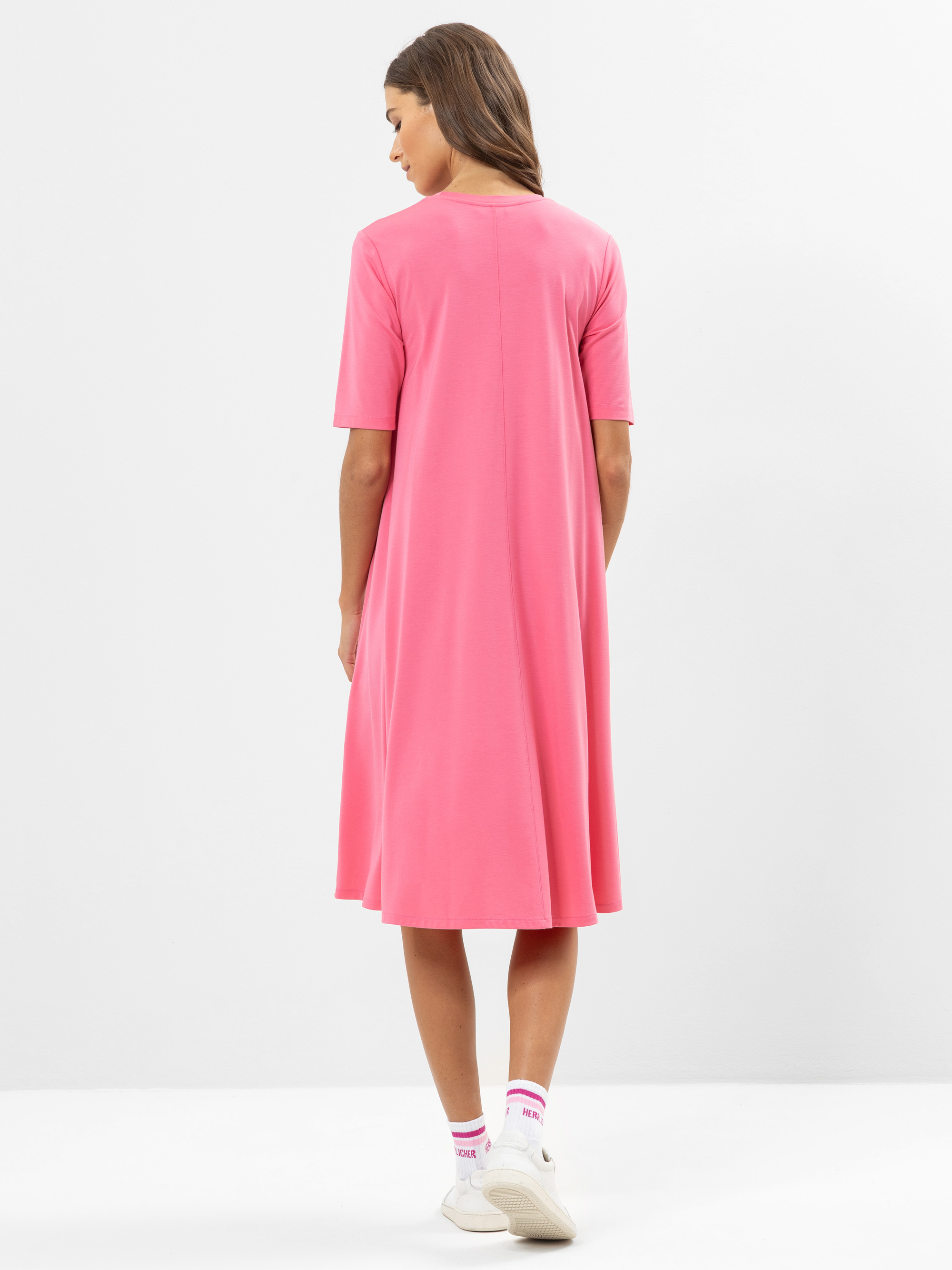 Eine Frau steht mit dem Gesicht nach hinten und trägt ein locker sitzendes, knielanges rosa Kleid mit kurzen Ärmeln. Sie hat langes braunes Haar und weiße Turnschuhe mit weißen Socken. Der Hintergrund ist einfarbig und hell.
