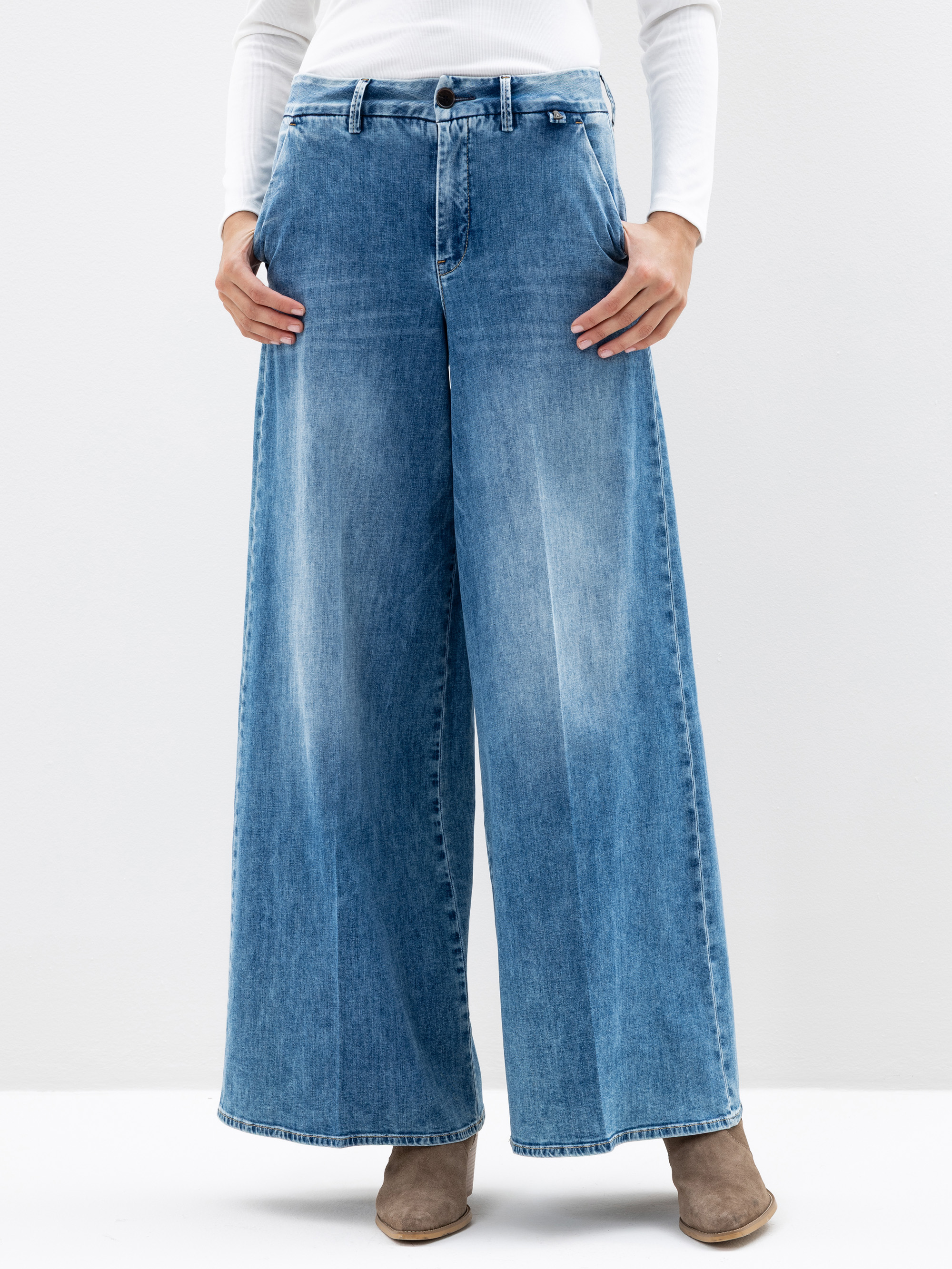 Eine Person, die eine blaue Jeans mit hoher Taille und weitem Bein, ein weißes langärmeliges Oberteil und taupefarbene Ankle Boots trägt, steht vor einem schlichten weißen Hintergrund. Das Foto ist knapp oberhalb der Schultern abgeschnitten.