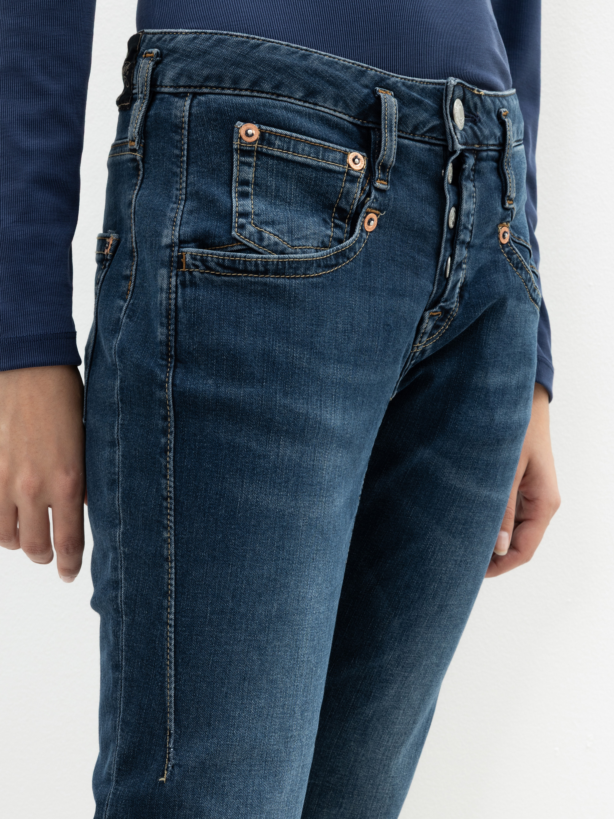 Eine Person, die dunkelblaue Jeans und ein langärmeliges marineblaues Oberteil trägt, steht vor einem schlichten weißen Hintergrund. Das Bild konzentriert sich auf den Jeansbund, die Vordertaschen und die Knopfdetails.