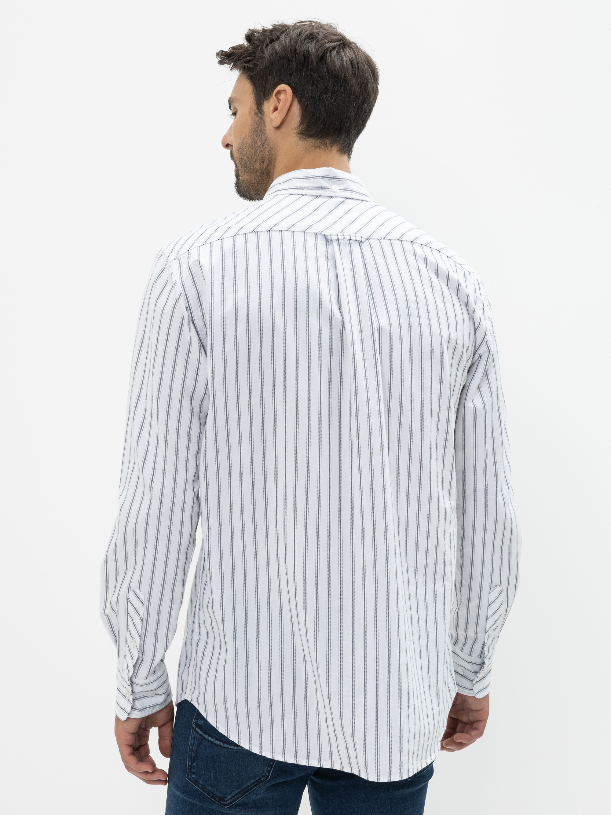 Ein Mann steht mit dem Rücken zur Kamera, trägt ein weißes Button-up-Hemd mit vertikalen schwarzen Streifen und eine blaue Jeans vor einem schlichten weißen Hintergrund.