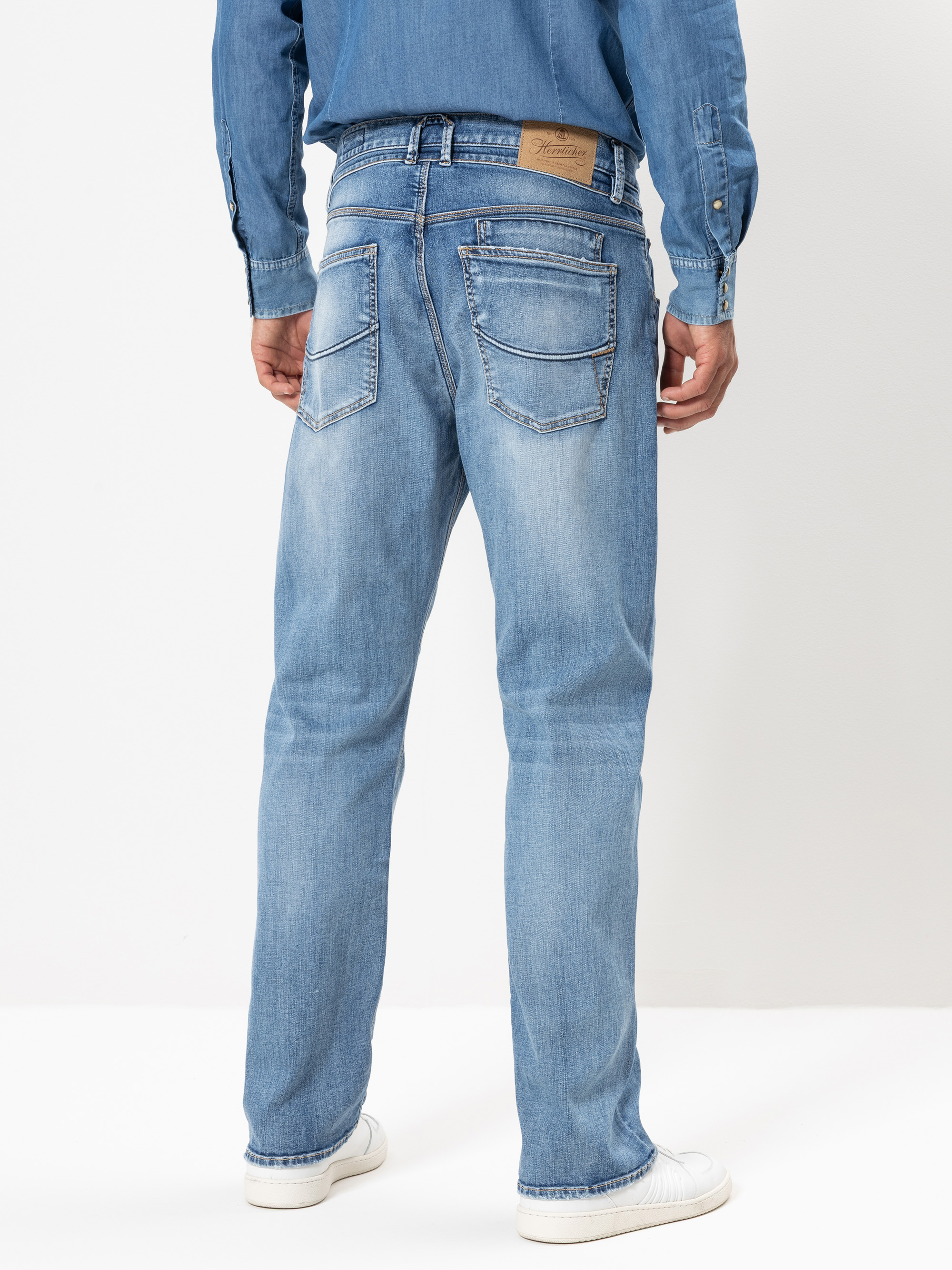Eine Person steht mit dem Gesicht nach hinten und trägt eine hellblaue Jeans, ein passendes blaues Jeanshemd und weiße Turnschuhe. Die Jeans hat sichtbare Nähte, Gesäßtaschen und einen hellbraunen Lederaufnäher am Bund.
