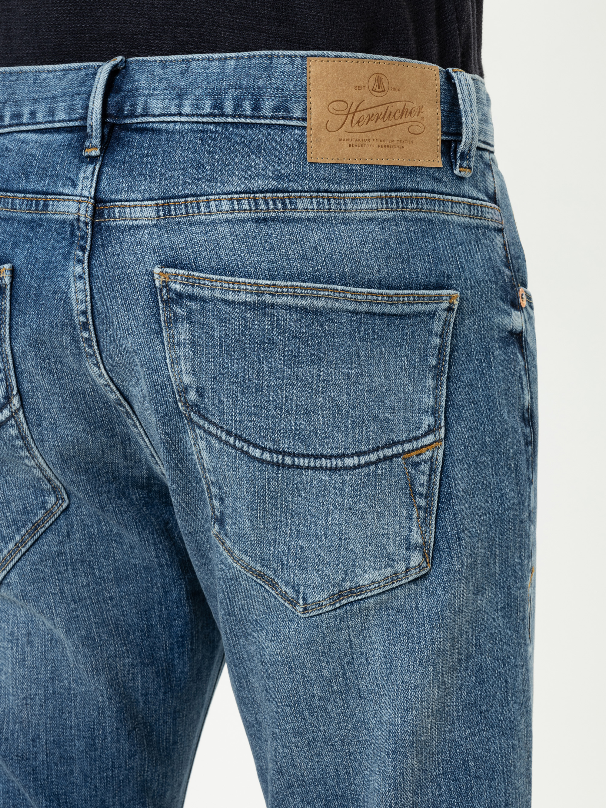 Nahaufnahme einer Person, die eine blaue Jeans trägt, mit der Gesäßtasche und einem braunen Lederaufnäher mit einem Markenlogo und Text in der Nähe des Hosenbundes. Der Hintergrund ist schlicht und hell.