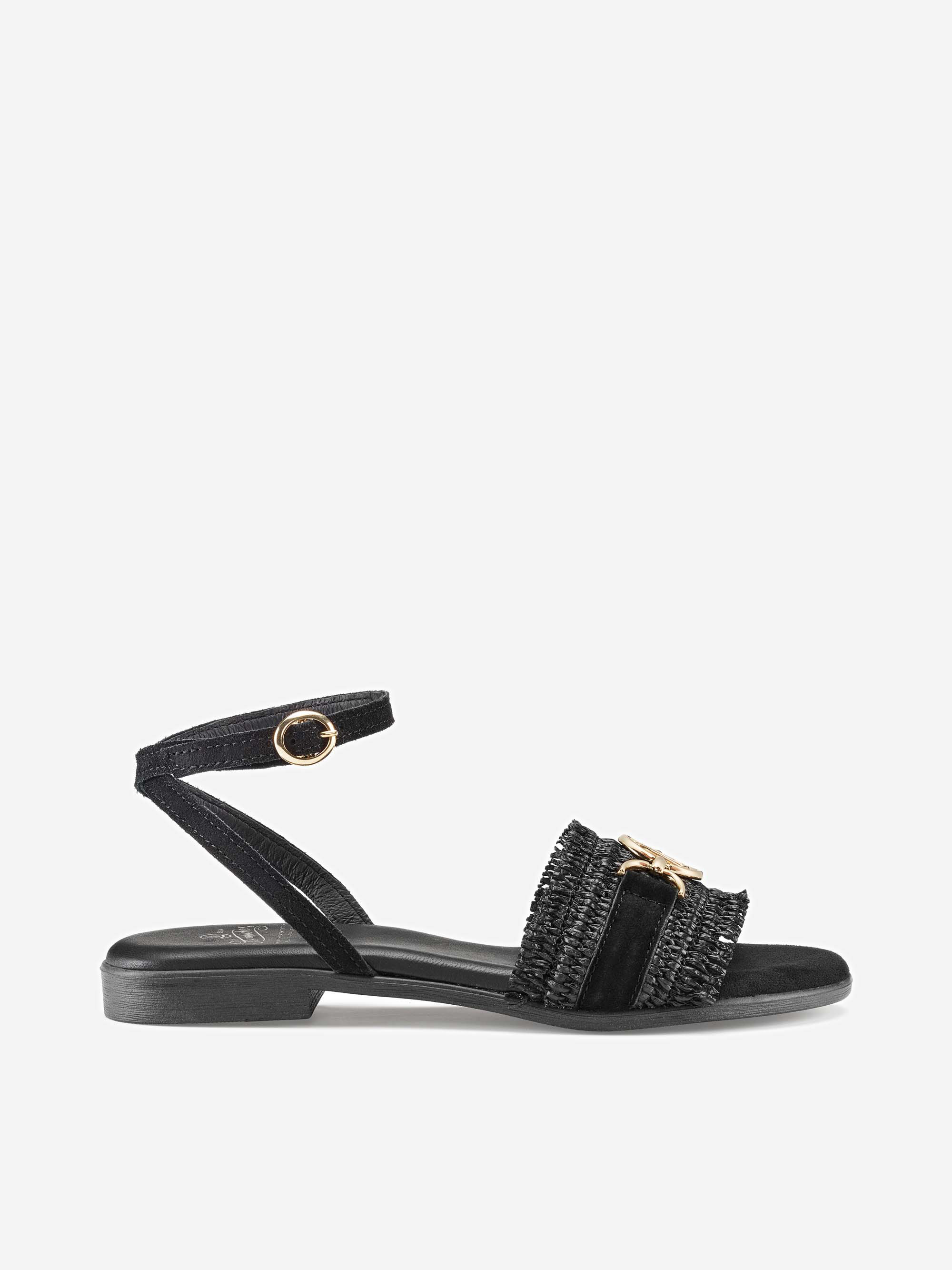 Eine schwarze flache Sandale mit Knöchelriemen und goldener Schnalle. Die Sandale hat einen strukturierten Vorderriemen mit dekorativen Details und einen niedrigen Absatz auf weißem Grund.