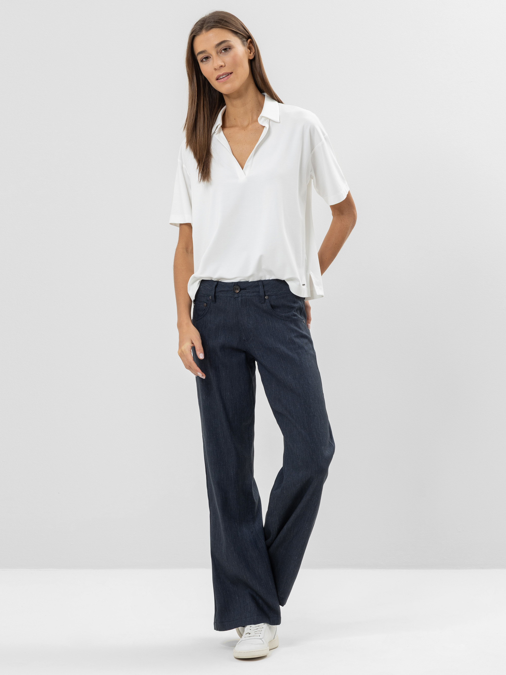 Eine Frau steht vor einem schlichten Hintergrund und trägt eine weiße kurzärmelige Bluse, dunkle Jeans mit weitem Bein und weiße Turnschuhe. Sie hat glattes braunes Haar und schaut mit entspanntem Blick in die Kamera.