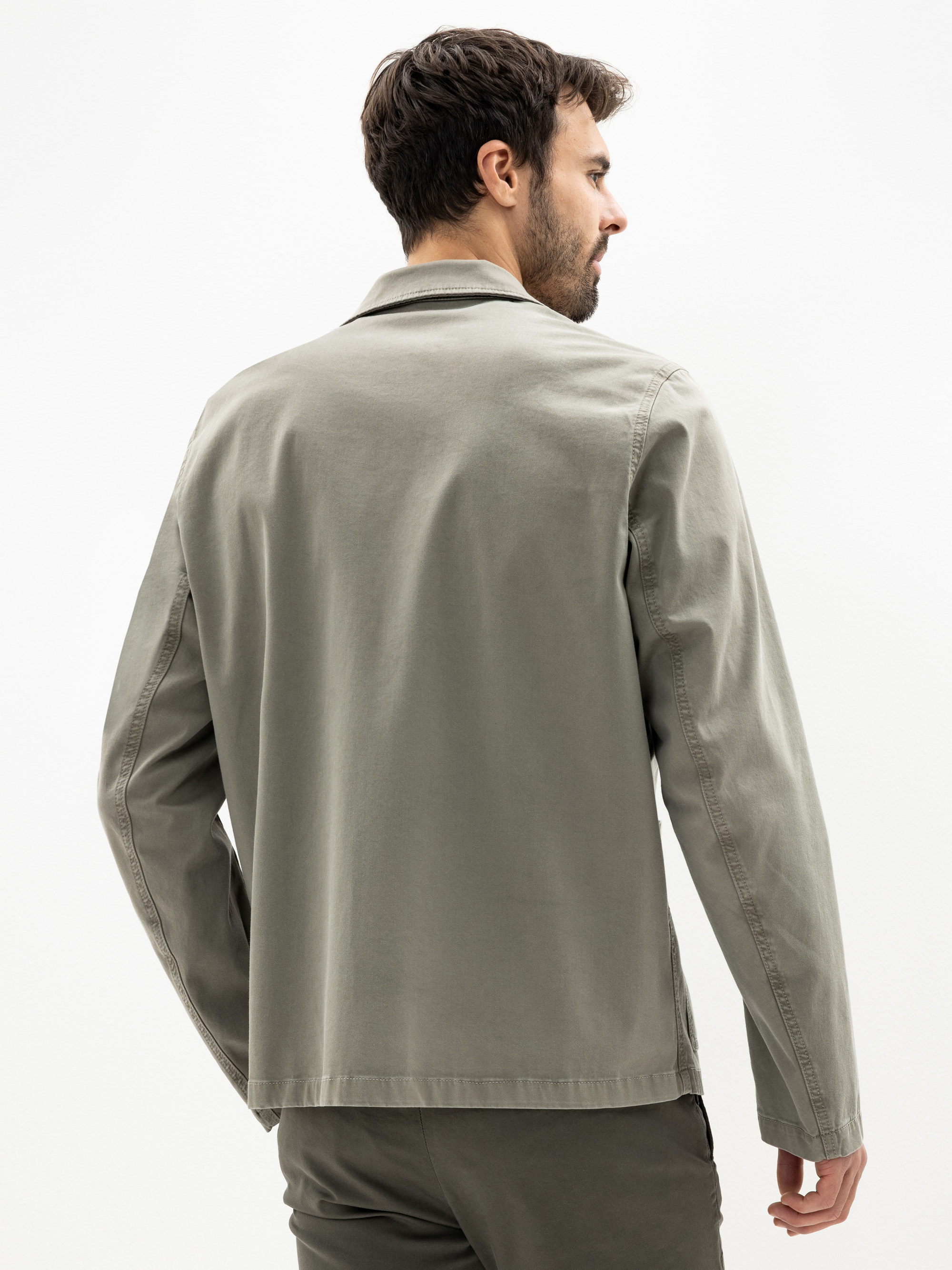 Ein Mann mit kurzen dunklen Haaren steht mit dem Gesicht zur Wand, trägt eine hellgraue Jacke und eine dazu passende Hose vor einem schlichten weißen Hintergrund.