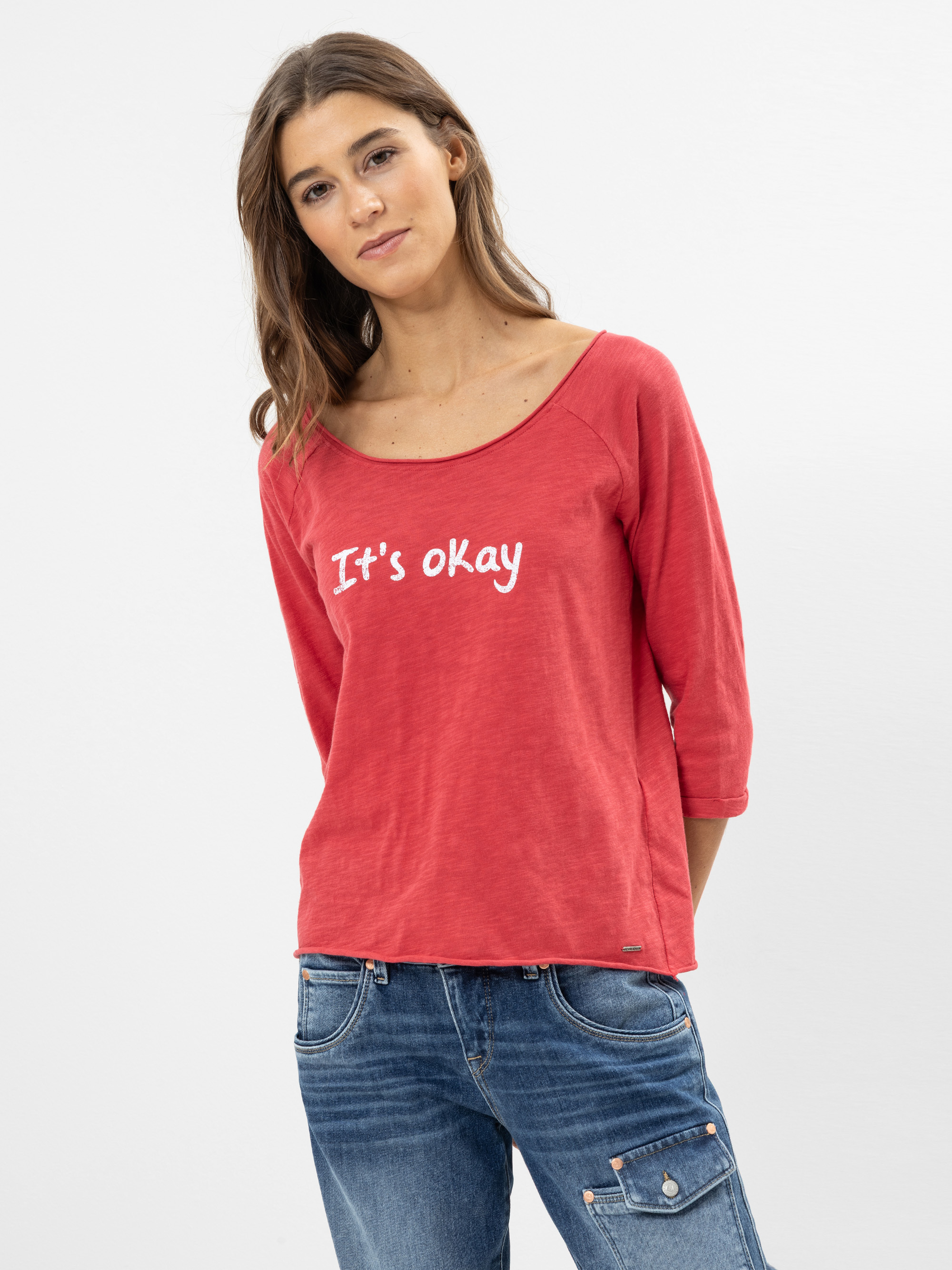 Eine Frau mit langen braunen Haaren trägt ein rotes Hemd mit dreiviertel Ärmeln und der Aufschrift Its okay in weiß sowie eine blaue Jeans. Sie steht vor einem schlichten, hellen Hintergrund und schaut mit neutralem Blick in die Kamera.