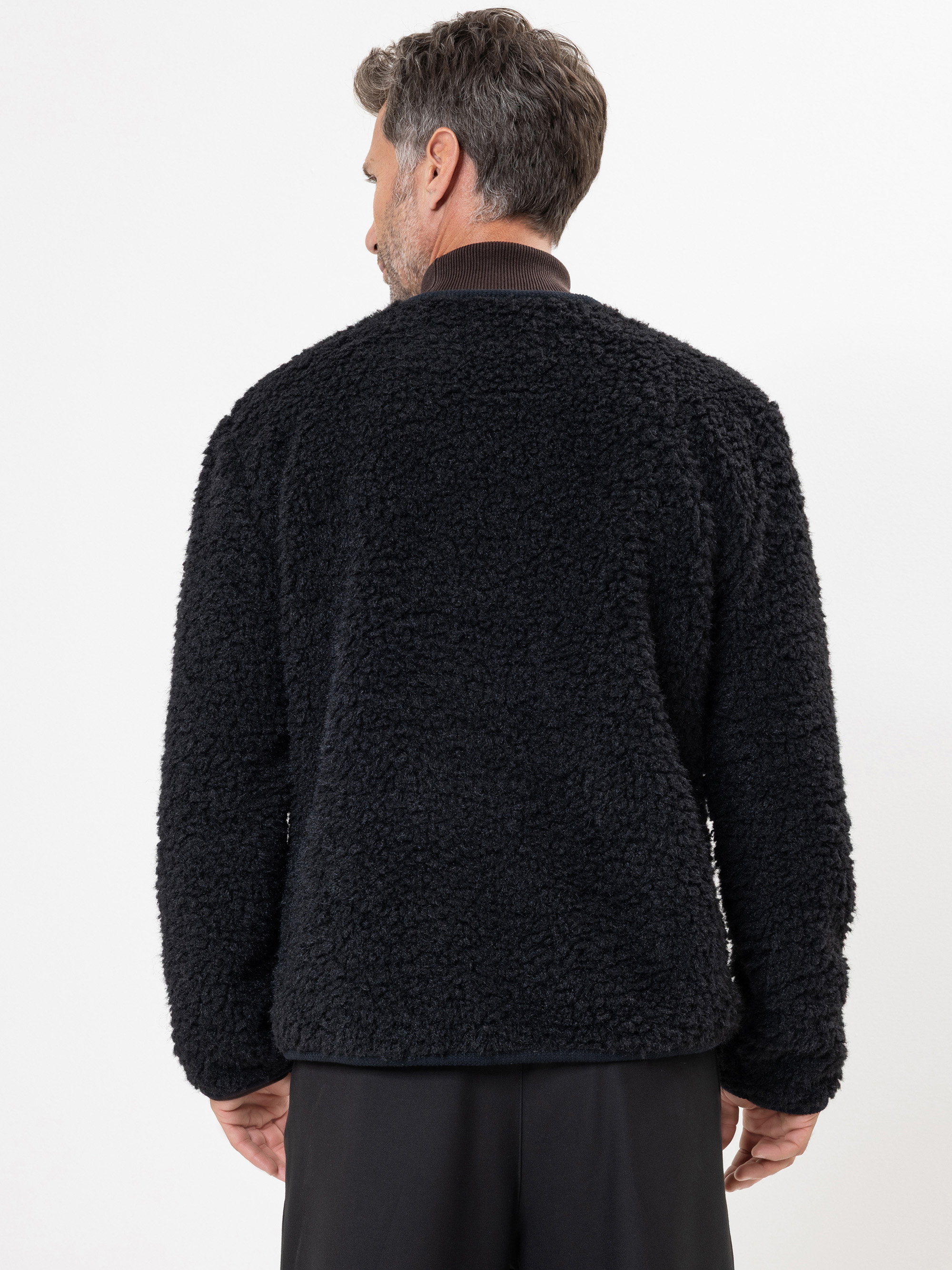Ein Mann mit kurzem, grauem Haar ist von hinten zu sehen, der eine schwarze, strukturierte Fleecejacke und eine schwarze Hose vor einem schlichten weißen Hintergrund trägt.