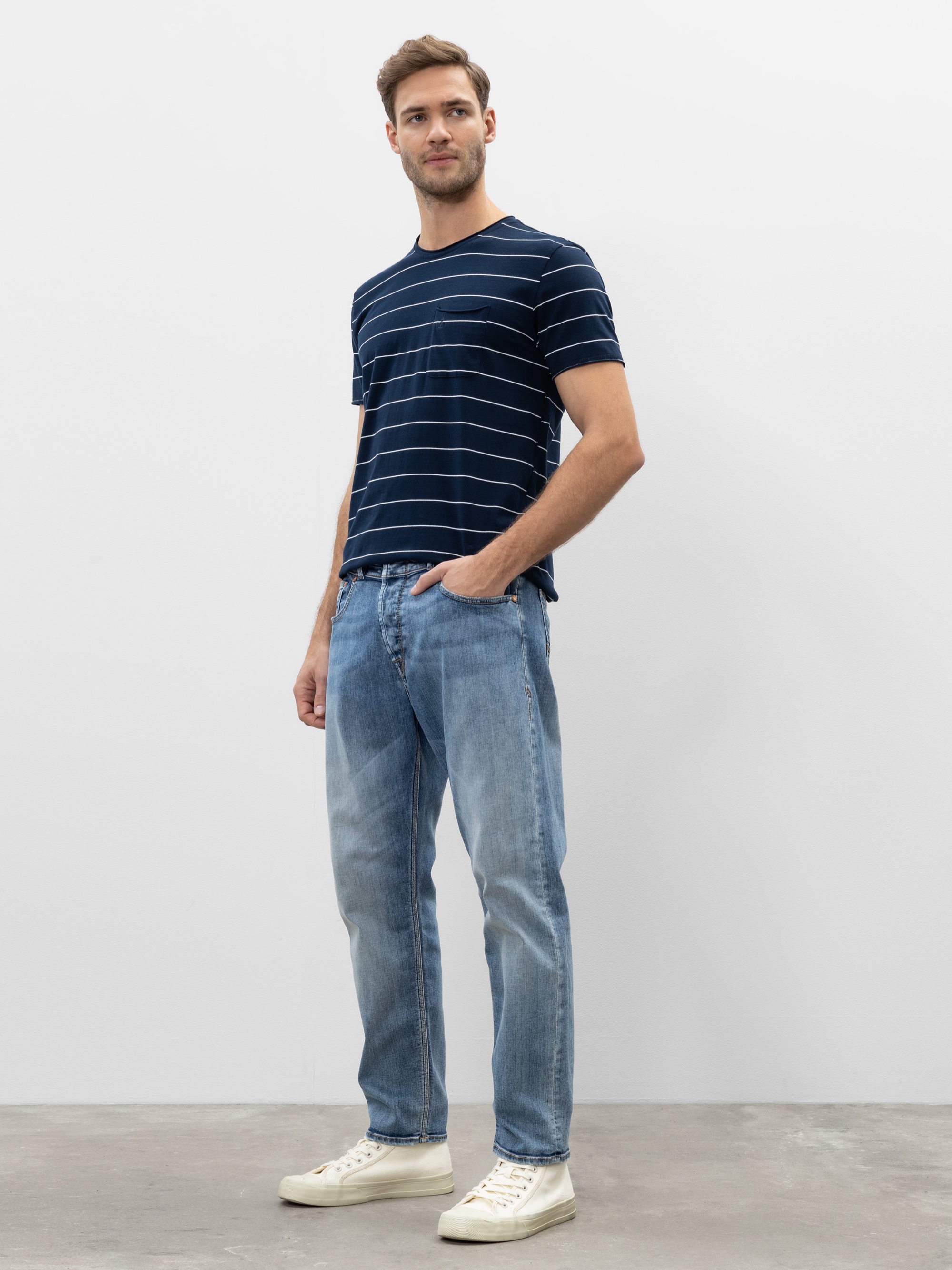 Ein Mann steht vor einer schlichten weißen Wand und trägt ein marineblau gestreiftes T-Shirt, hellblaue Jeans und weiße Turnschuhe. Er hat eine Hand in der Tasche und blickt leicht zur Seite.
