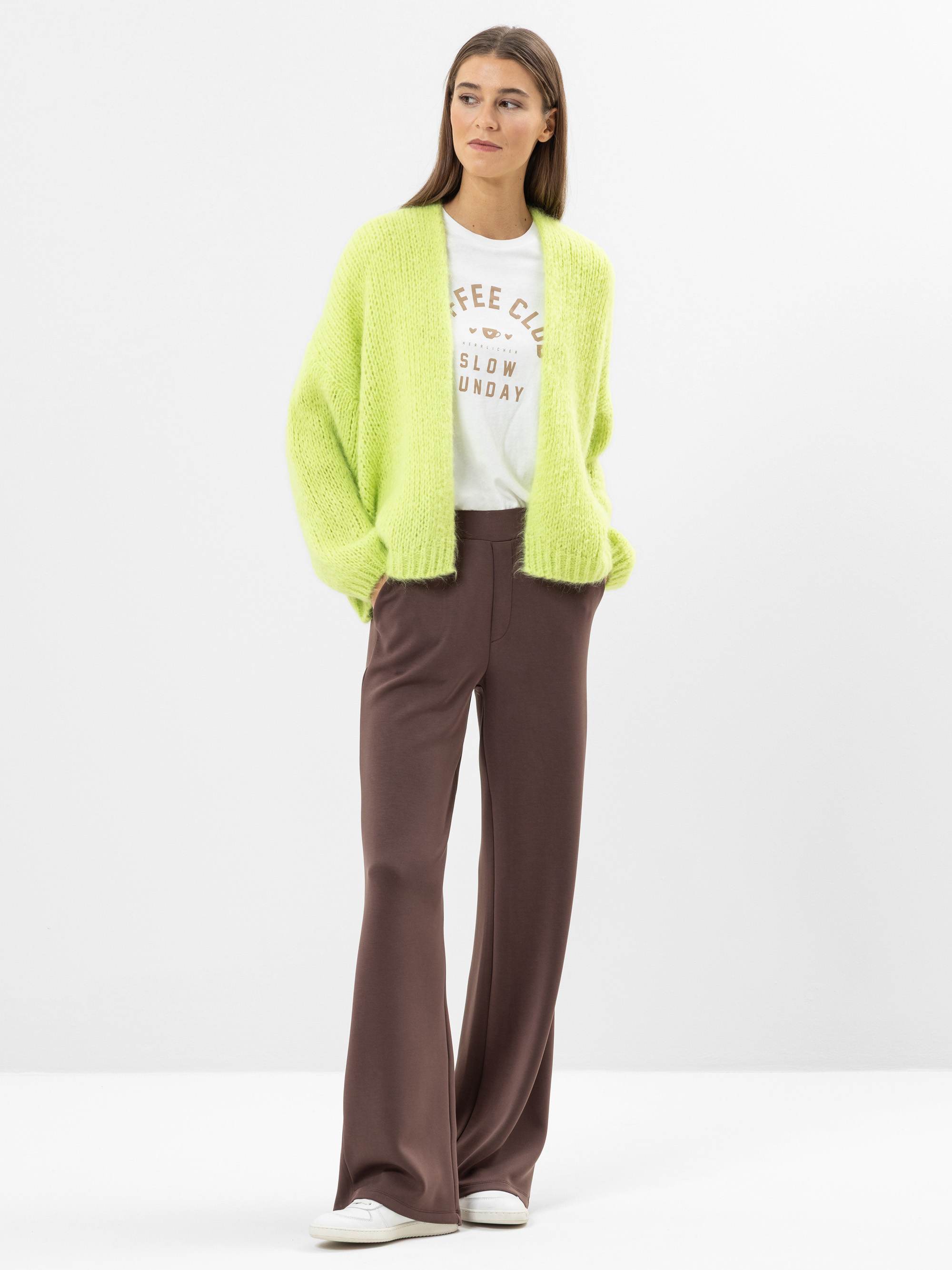 Eine Frau steht vor einem weißen Hintergrund und trägt eine hellgrüne Strickjacke, ein weißes bedrucktes T-Shirt, eine braune Hose mit weitem Bein und weiße Turnschuhe. Sie hat langes braunes Haar und einen neutralen Gesichtsausdruck.
