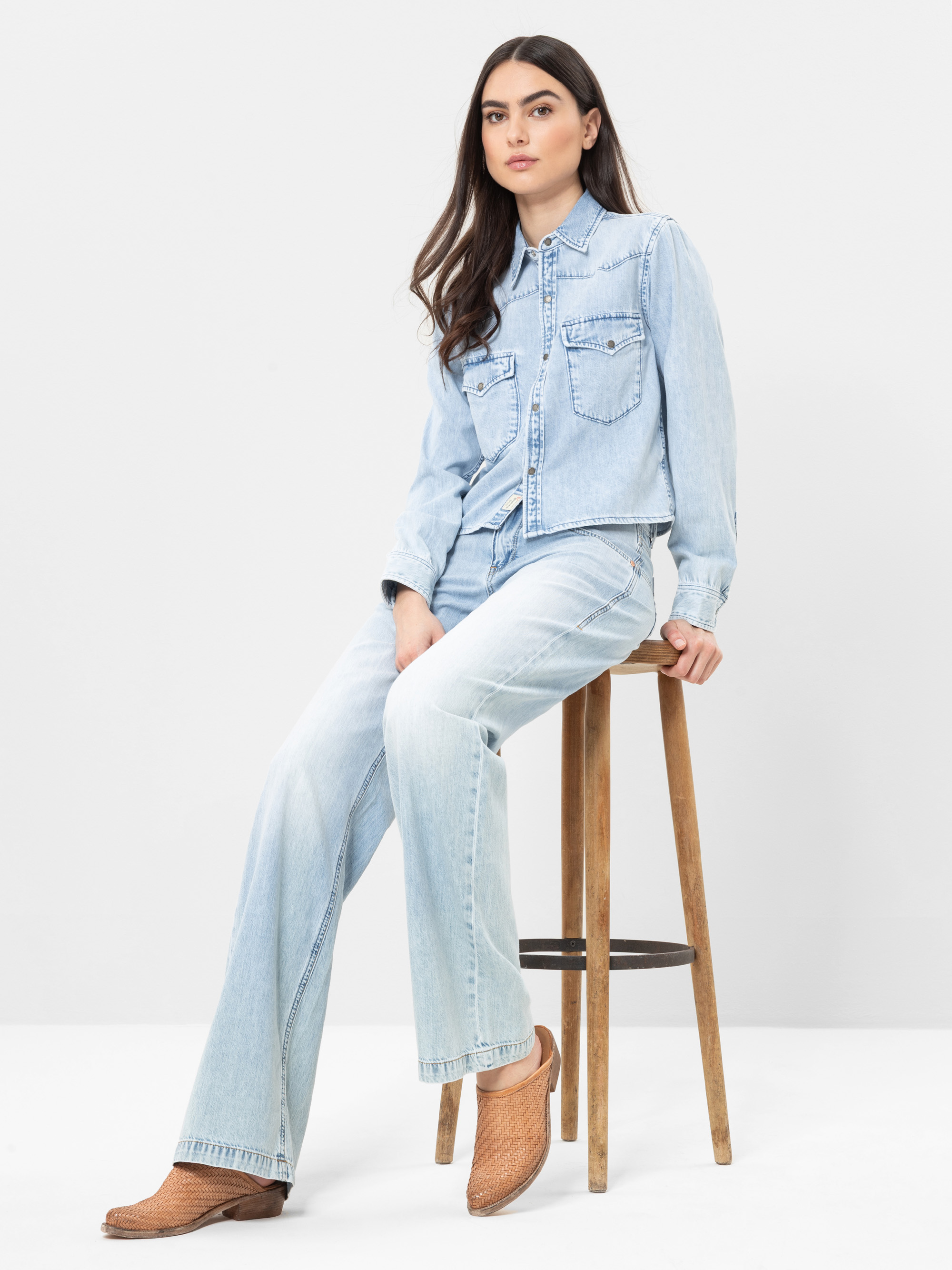 Eine Frau mit langen dunklen Haaren sitzt auf einem Holzschemel, trägt ein hellblaues Jeanshemd und Jeans mit braunen gewebten Schuhen. Sie blickt mit neutralem Gesichtsausdruck vor einem schlichten weißen Hintergrund nach vorne.