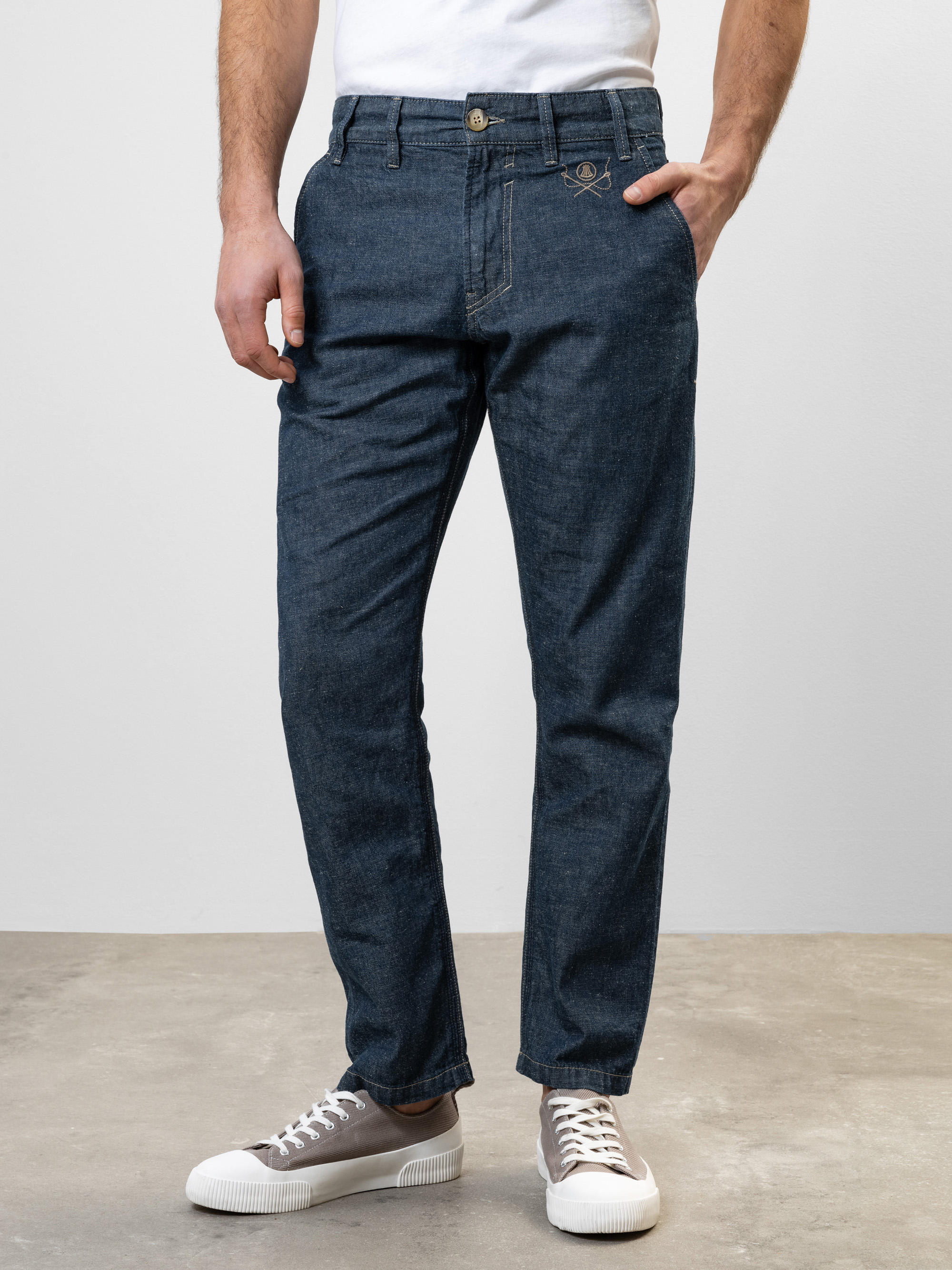Ein Mann in dunkelblauen Jeans, weißem Hemd und grauen Turnschuhen steht mit einer Hand in der Tasche auf einem hellen Boden vor einer schlichten hellgrauen Wand.