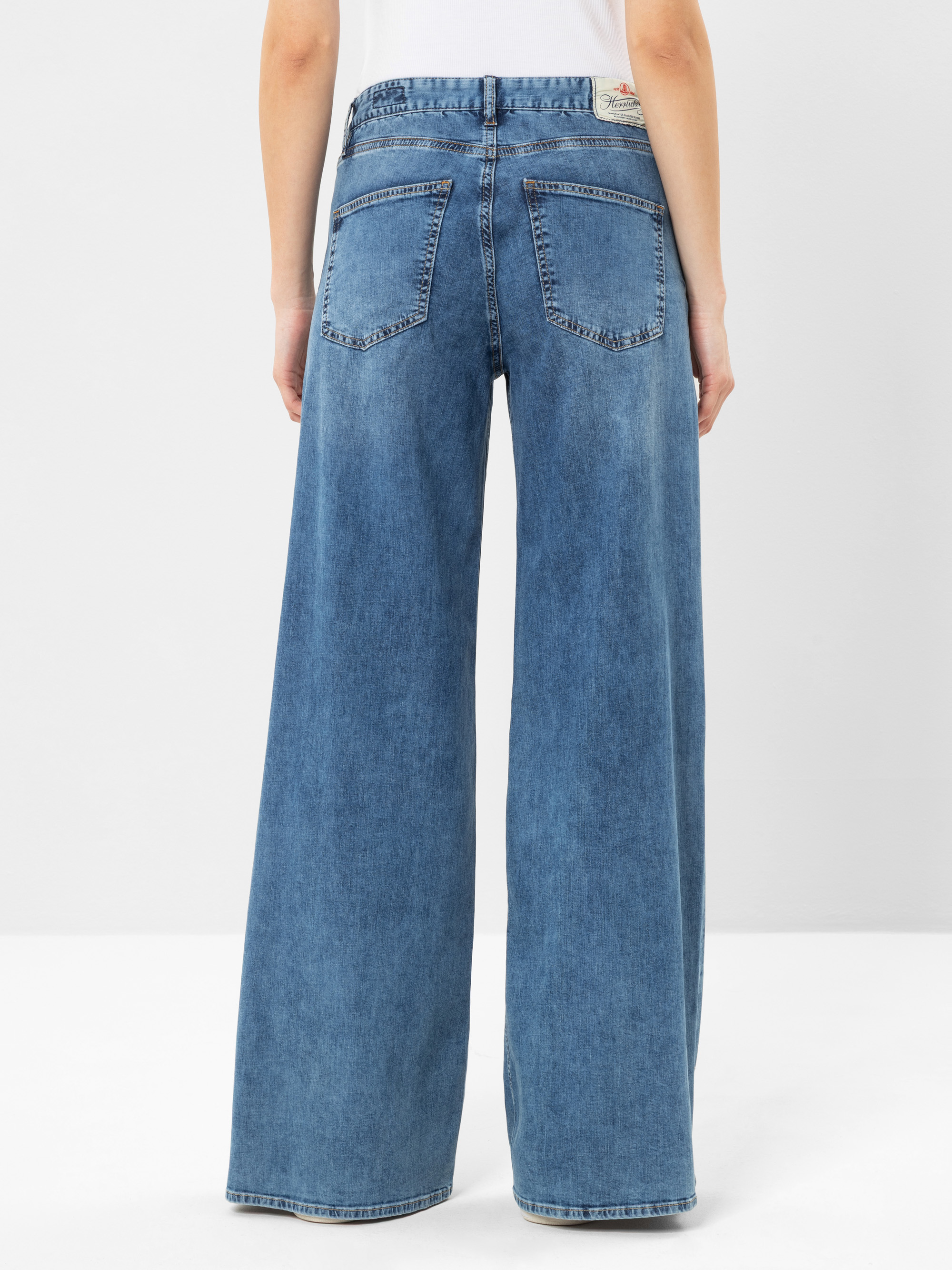 Eine Person trägt eine hellblaue Jeans mit hoher Taille und weitem Bein von hinten gesehen. Die Jeans hat zwei Gesäßtaschen und ein kleines hellbraunes Etikett am Bund. Der Hintergrund ist einfarbig weiß.