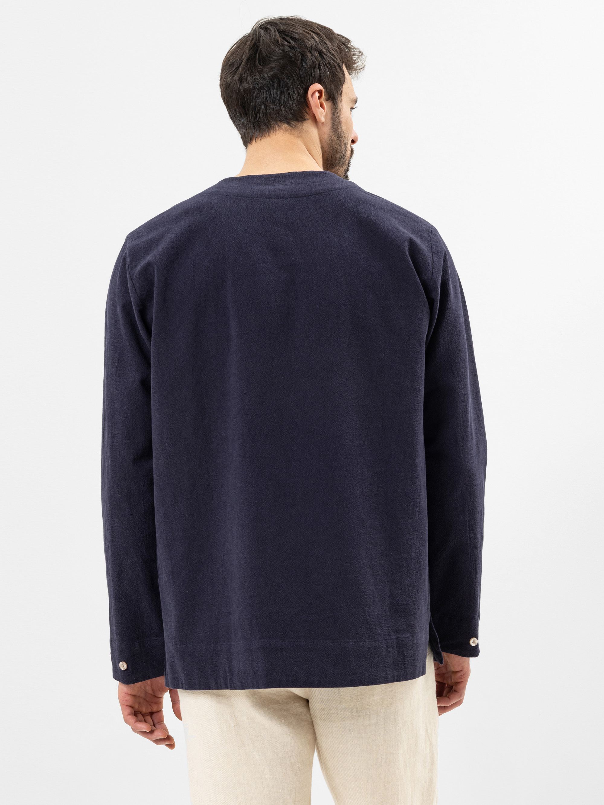 Ein Mann steht von der Kamera abgewandt, trägt ein dunkles, marineblaues, langärmeliges Hemd und eine hellbeige Hose vor einem schlichten weißen Hintergrund.