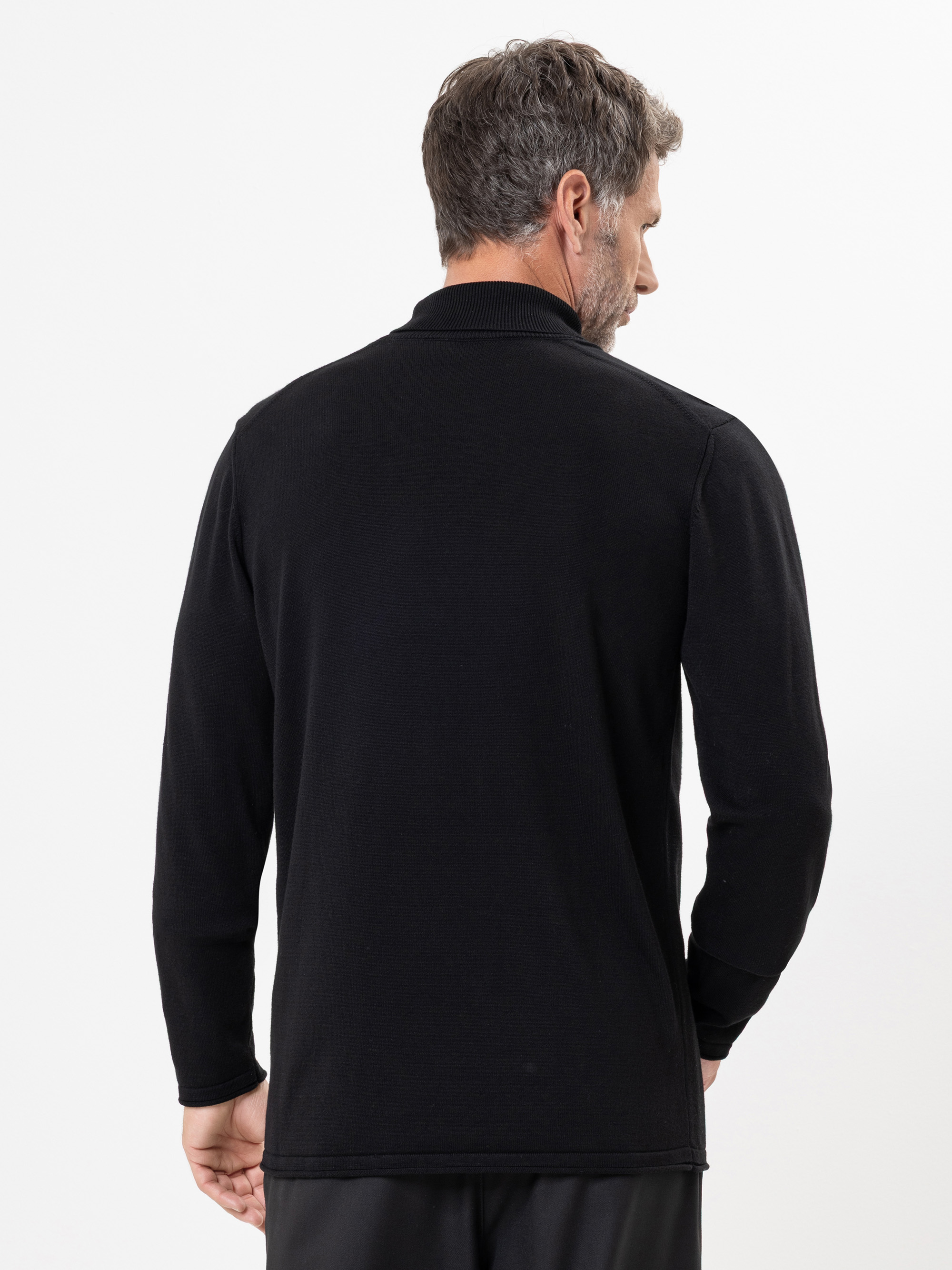 Ein Mann mit kurzen braunen Haaren wird von hinten gezeigt. Er trägt ein schwarzes langärmeliges Polohemd und eine schwarze Hose vor einem schlichten weißen Hintergrund.
