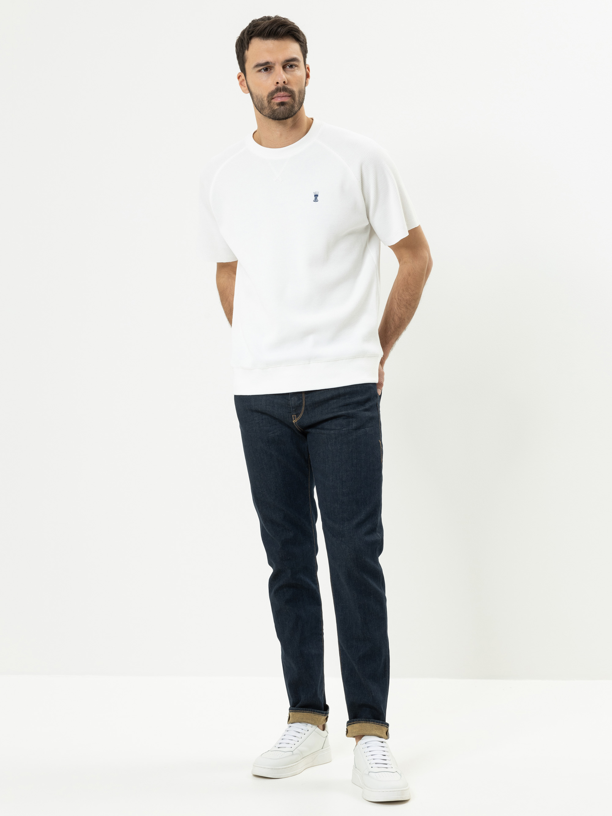 Ein Mann steht vor einem schlichten weißen Hintergrund und trägt ein kurzärmeliges weißes T-Shirt, dunkelblaue Jeans und weiße Turnschuhe. Er hat die Hände hinter dem Rücken verschränkt und blickt leicht zur Seite.