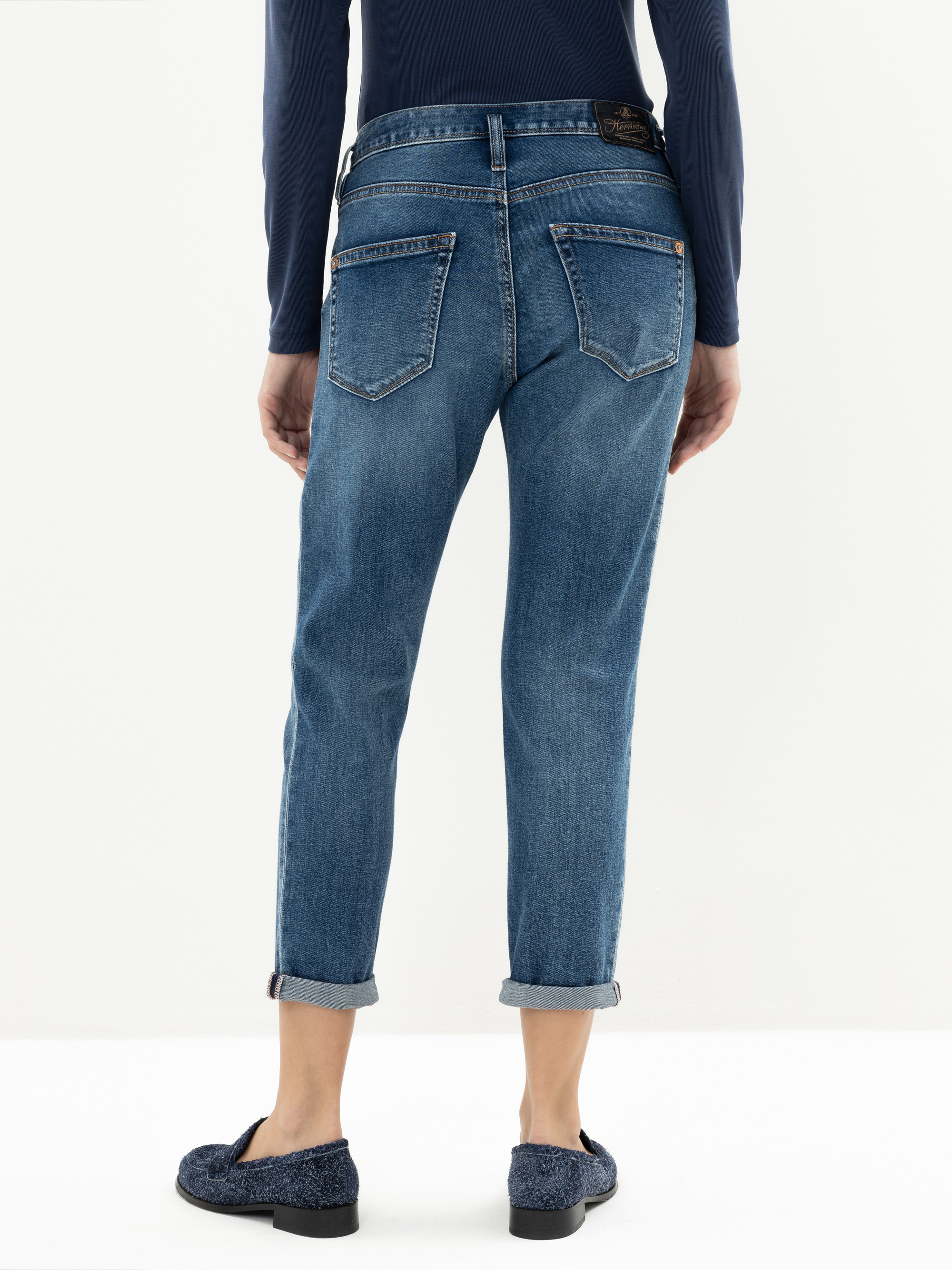 Eine Person in blauen Jeans mit Manschetten und marineblauen Halbschuhen steht mit dem Rücken zur Kamera. Die Person trägt außerdem ein langärmeliges marineblaues Oberteil.