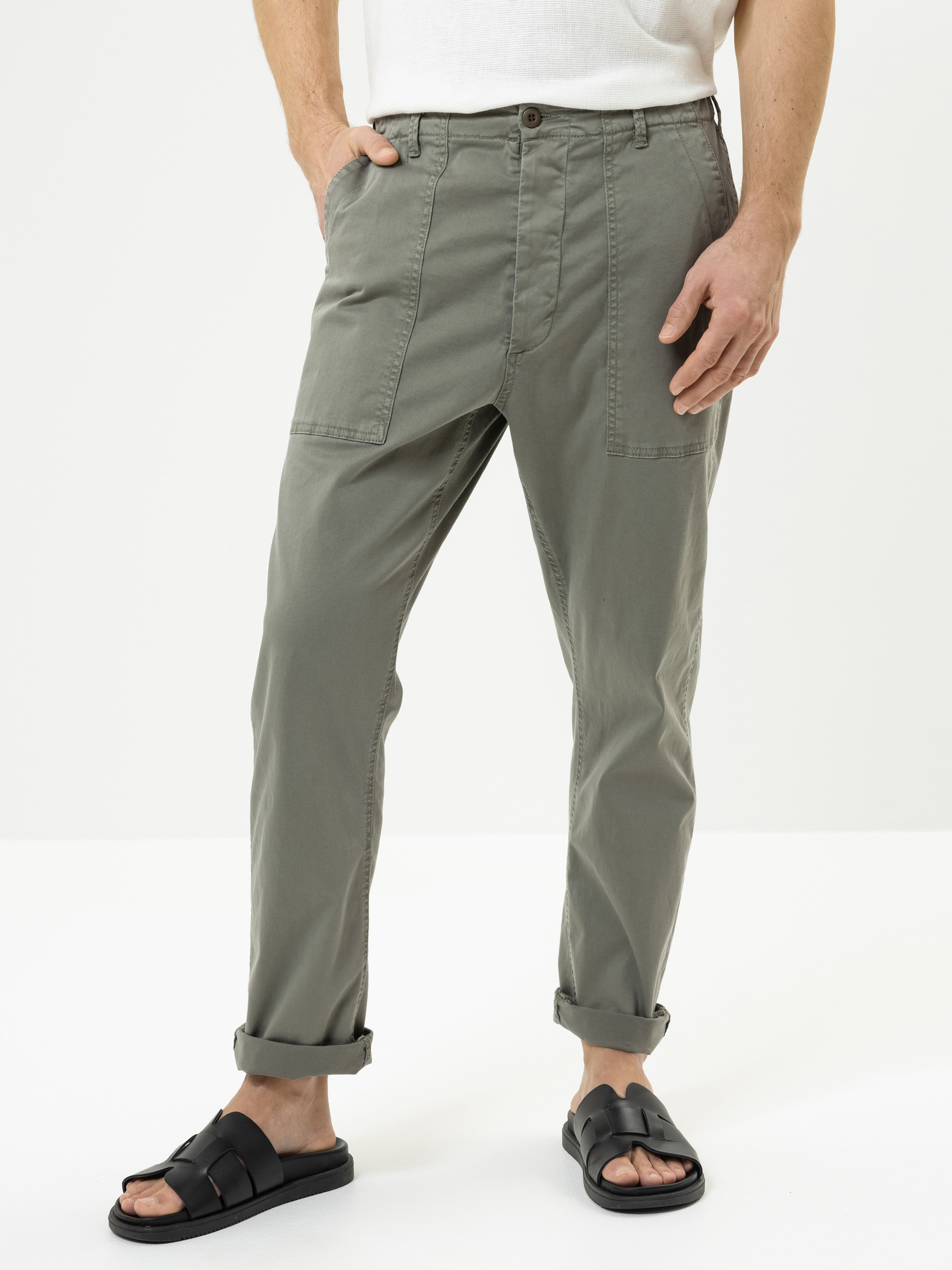 Eine Person, die eine olivgrüne Hose mit großen Vordertaschen, ein weißes Hemd, schwarze Sandalen und aufgerollte Manschetten trägt, steht mit den Händen in den Taschen vor einem schlichten weißen Hintergrund.