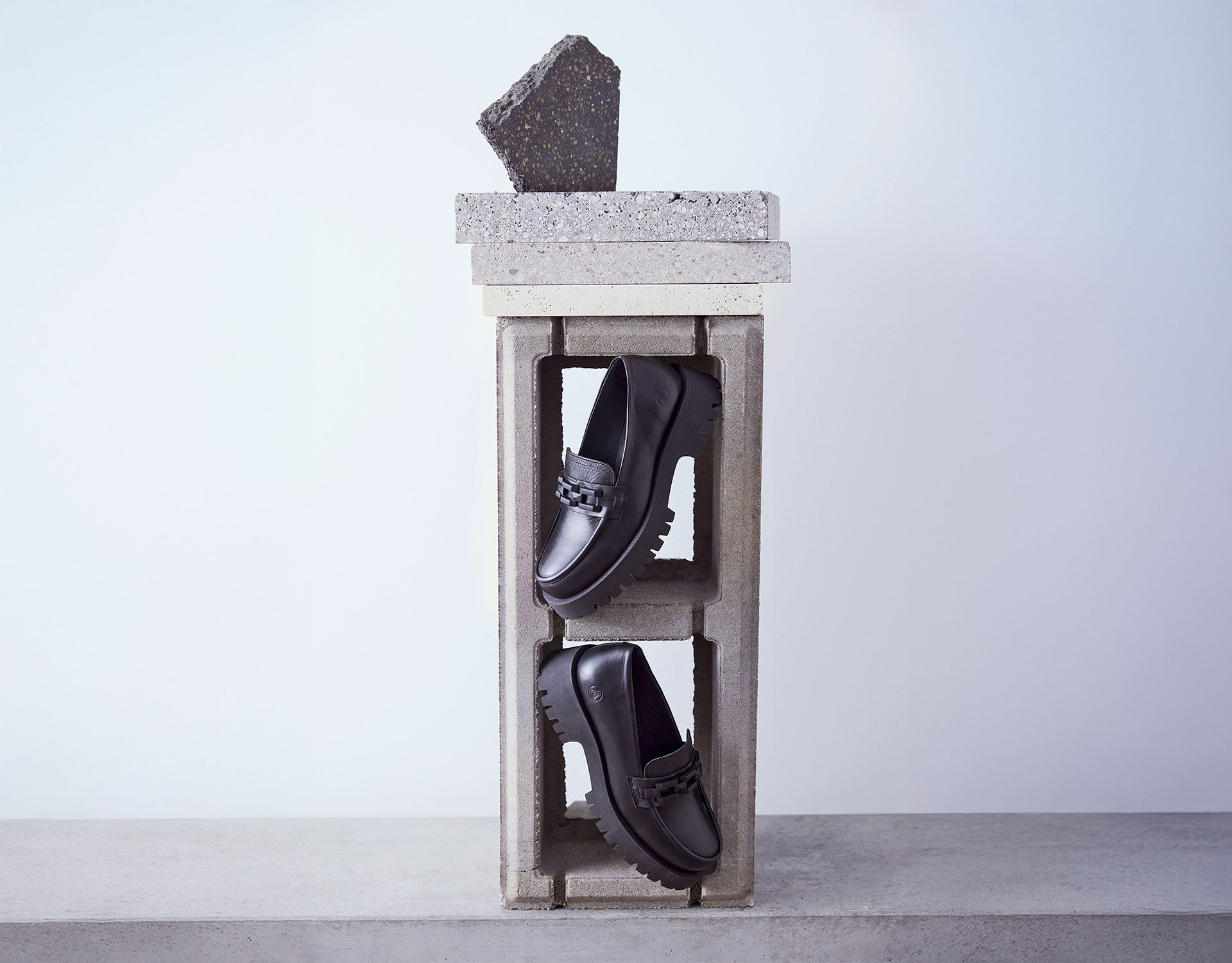 Zwei schwarze Halbschuhe, die senkrecht in einem hohlen Betonblock stehen, auf dem weitere Betonblöcke und ein dunkler Stein gestapelt sind, vor einem schlichten hellen Hintergrund.