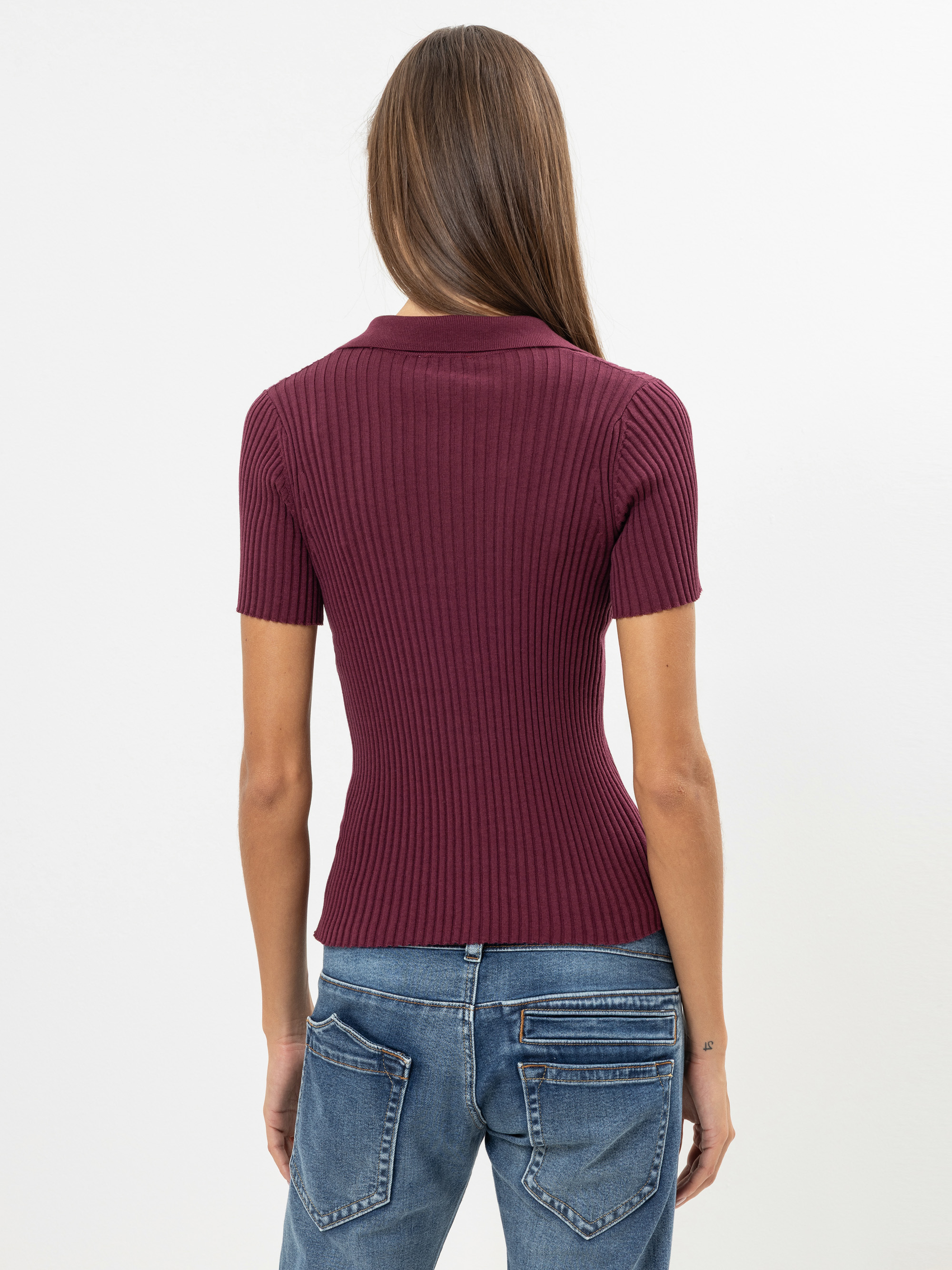 Eine Frau mit langen braunen Haaren ist von hinten zu sehen. Sie trägt ein kurzärmeliges, geripptes kastanienbraunes Oberteil und blaue Jeans vor einem schlichten weißen Hintergrund.