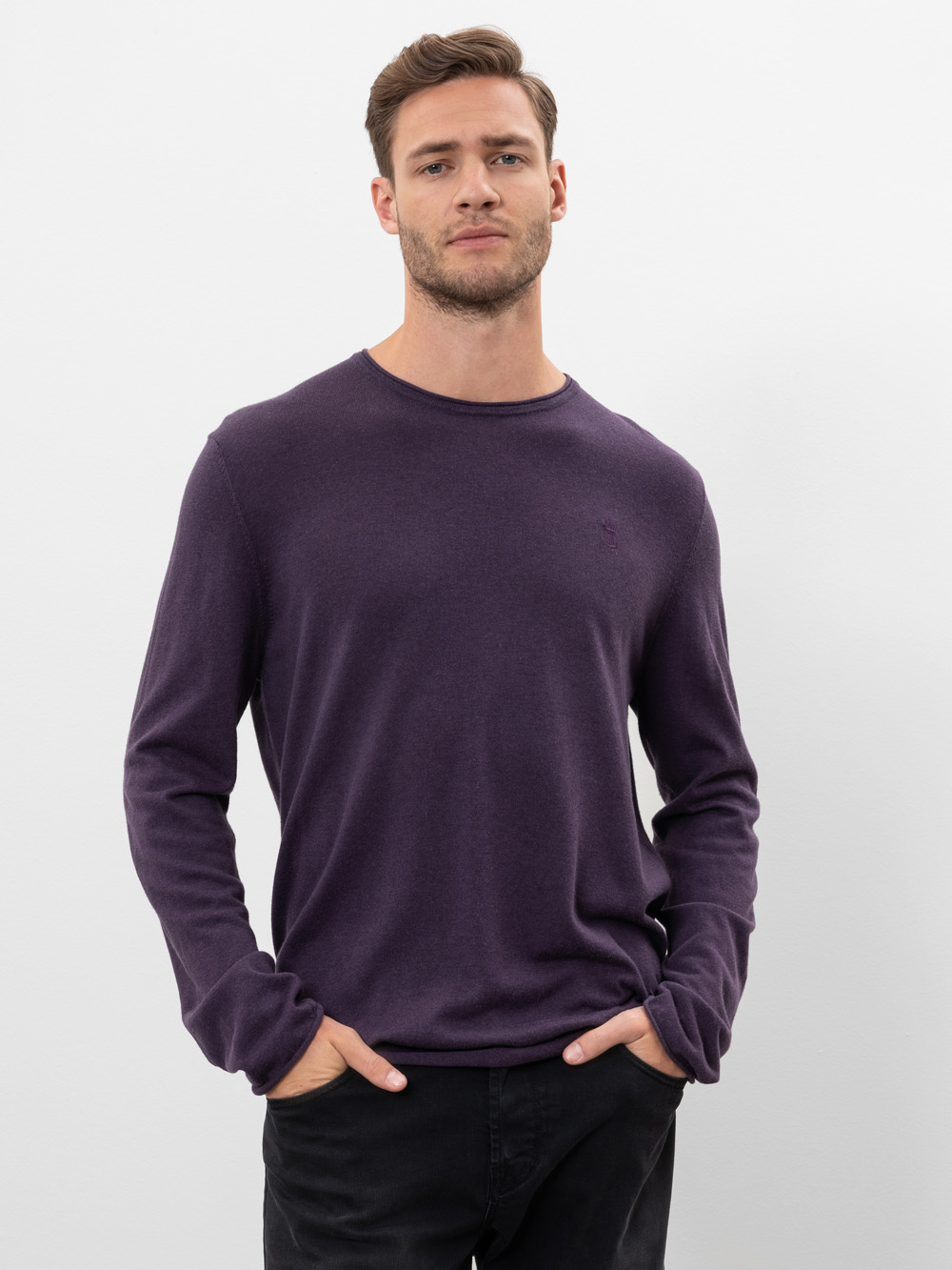 Ein Mann mit kurzem braunem Haar und gestutztem Bart trägt einen langärmeligen, dunkelvioletten Pullover und eine schwarze Hose und steht mit den Händen in den Taschen vor einem schlichten weißen Hintergrund.