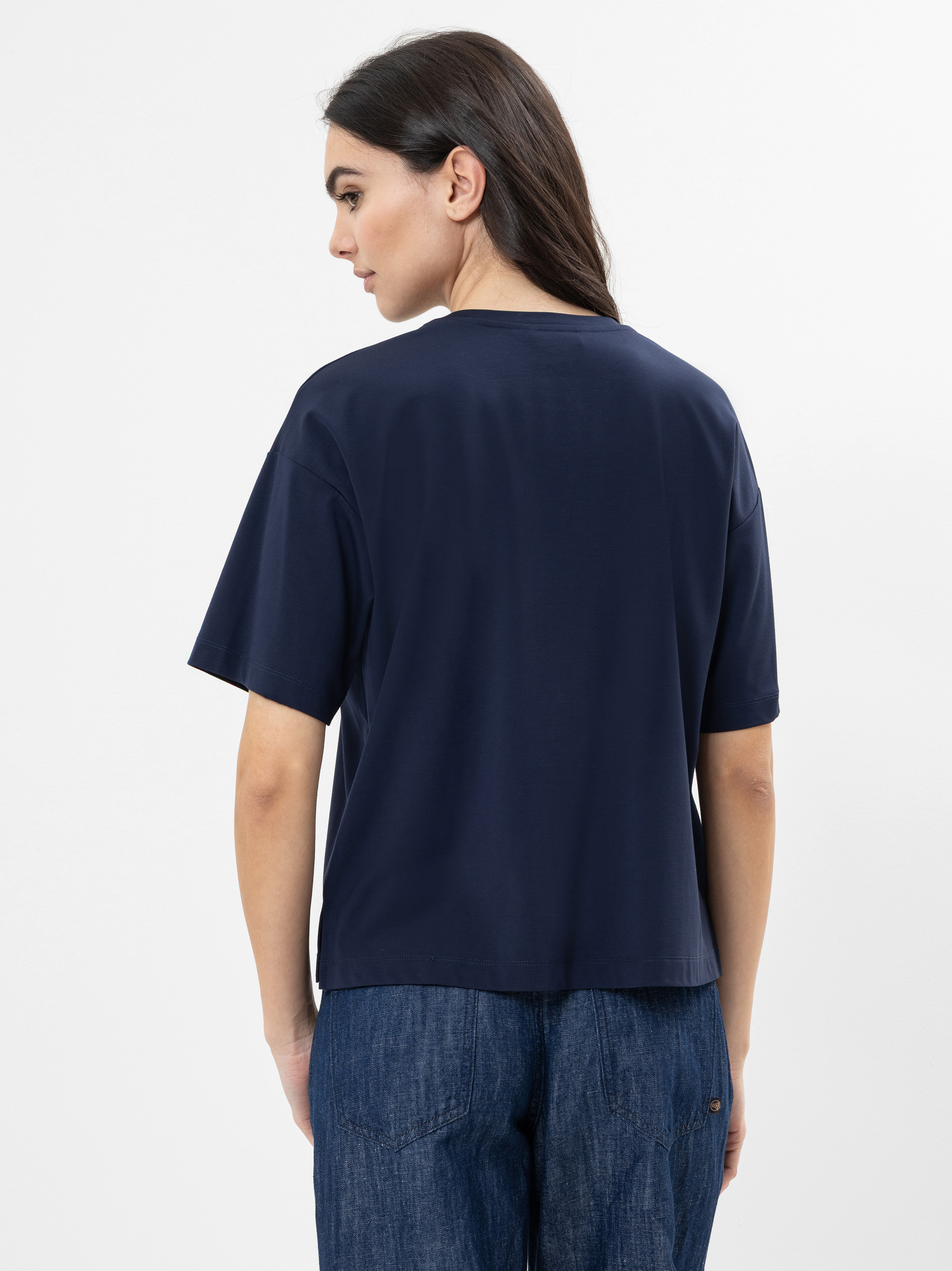 Man sieht eine Person mit langen dunklen Haaren von hinten, die ein locker sitzendes marineblaues T-Shirt und eine blaue Jeans vor einem schlichten weißen Hintergrund trägt.