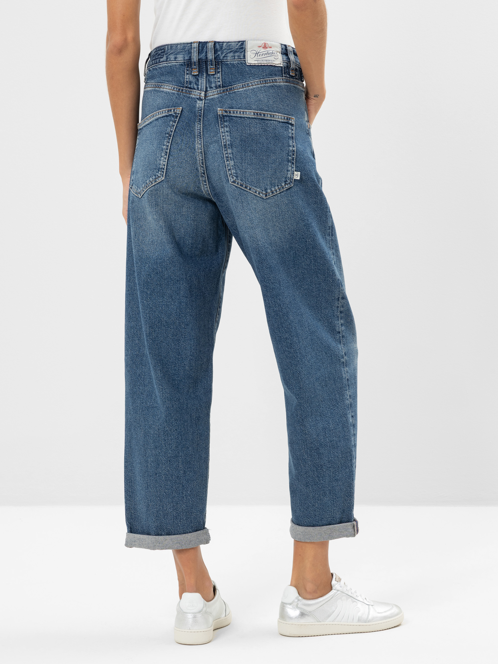 Person, die vor einem schlichten Hintergrund steht und eine aufgerollte blaue Jeans, ein weißes Hemd und weiße Turnschuhe trägt. Das Bild ist von hinten aufgenommen und konzentriert sich auf die Jeans und die Schuhe.