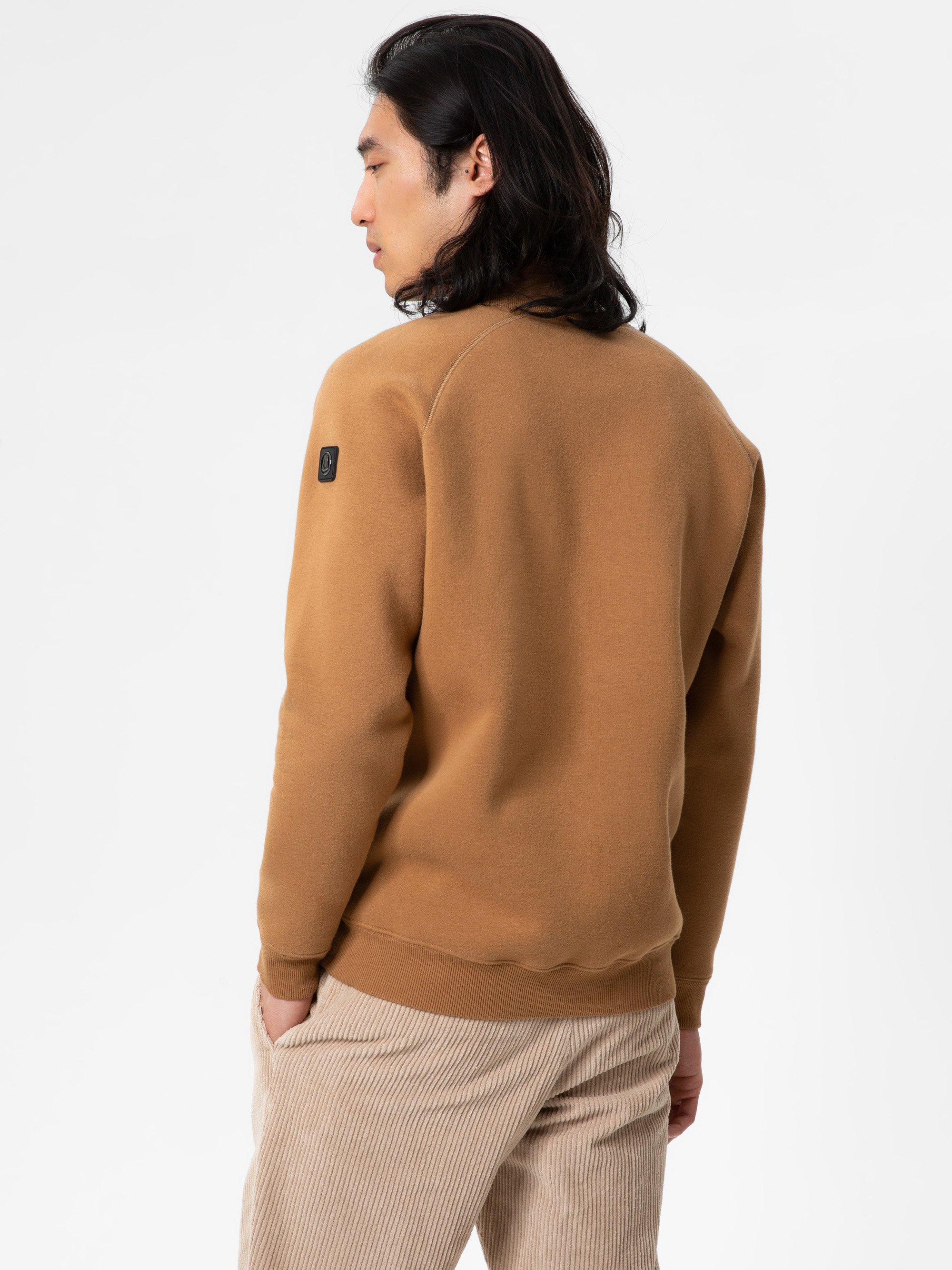 Eine Person mit langen dunklen Haaren, die ein kamelbraunes Sweatshirt und eine beigefarbene Cordhose trägt, steht mit einer Hand in der Hosentasche vor einem schlichten weißen Hintergrund und wendet sich von der Kamera ab.