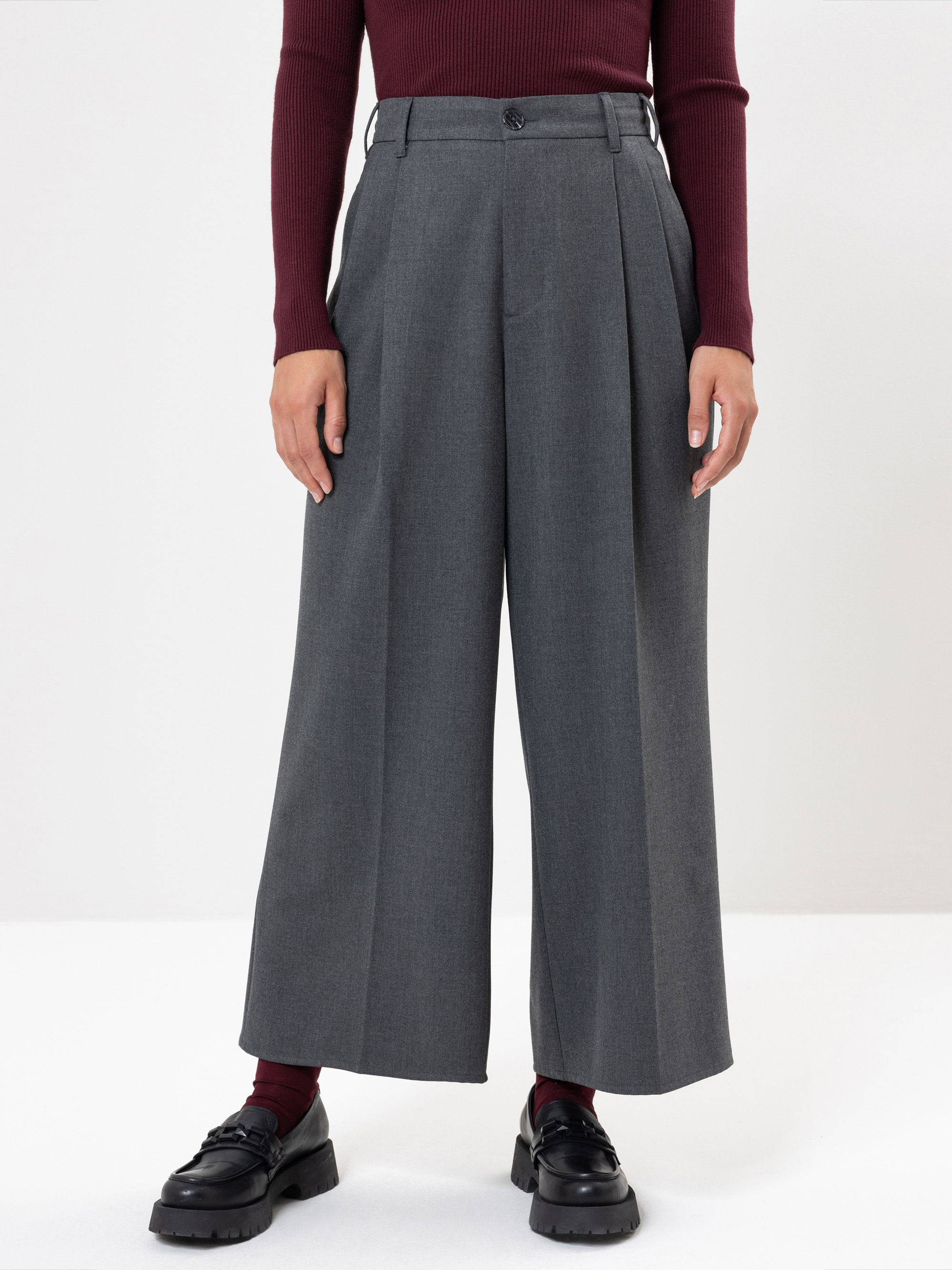 Eine Person, die eine graue Hose mit hoher Taille und weitem Bein mit Bügelfalten, ein weinrotes, langärmeliges Rippenoberteil, weinrote Socken und schwarze, klobige Slipper trägt, steht vor einem schlichten weißen Hintergrund.