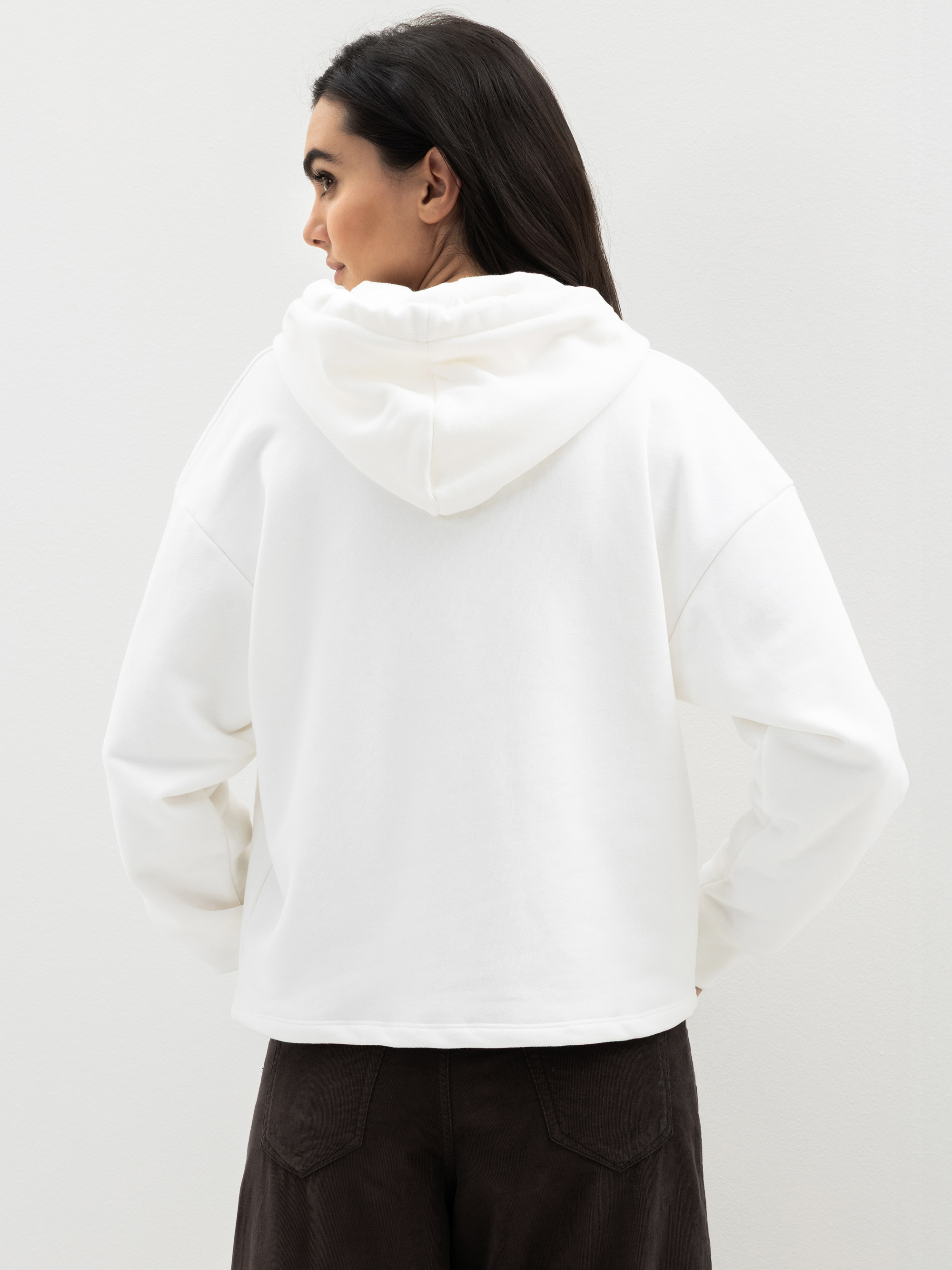 Eine Frau mit langen dunklen Haaren steht mit dem Gesicht zur Wand, trägt ein weißes Sweatshirt mit Kapuze und eine dunkle Hose und steht vor einem schlichten hellen Hintergrund.