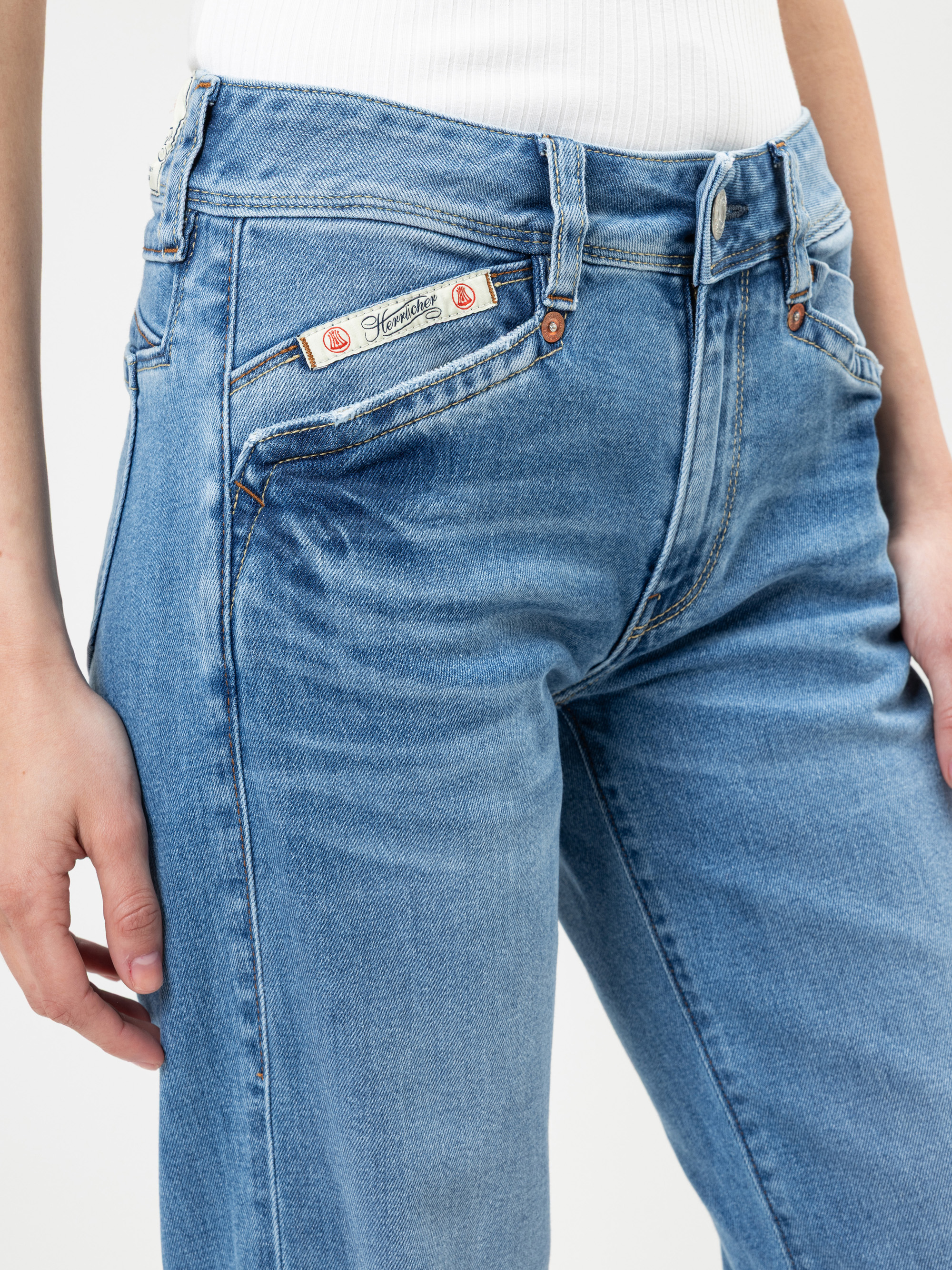 Eine Person trägt eine hellblaue Jeans mit einem Aufnäher mit der Aufschrift "Stay Loose" über der Tasche und ein weißes geripptes Oberteil. Nur der Unterkörper und die Oberschenkel sind vor einem einfarbigen Hintergrund zu sehen.