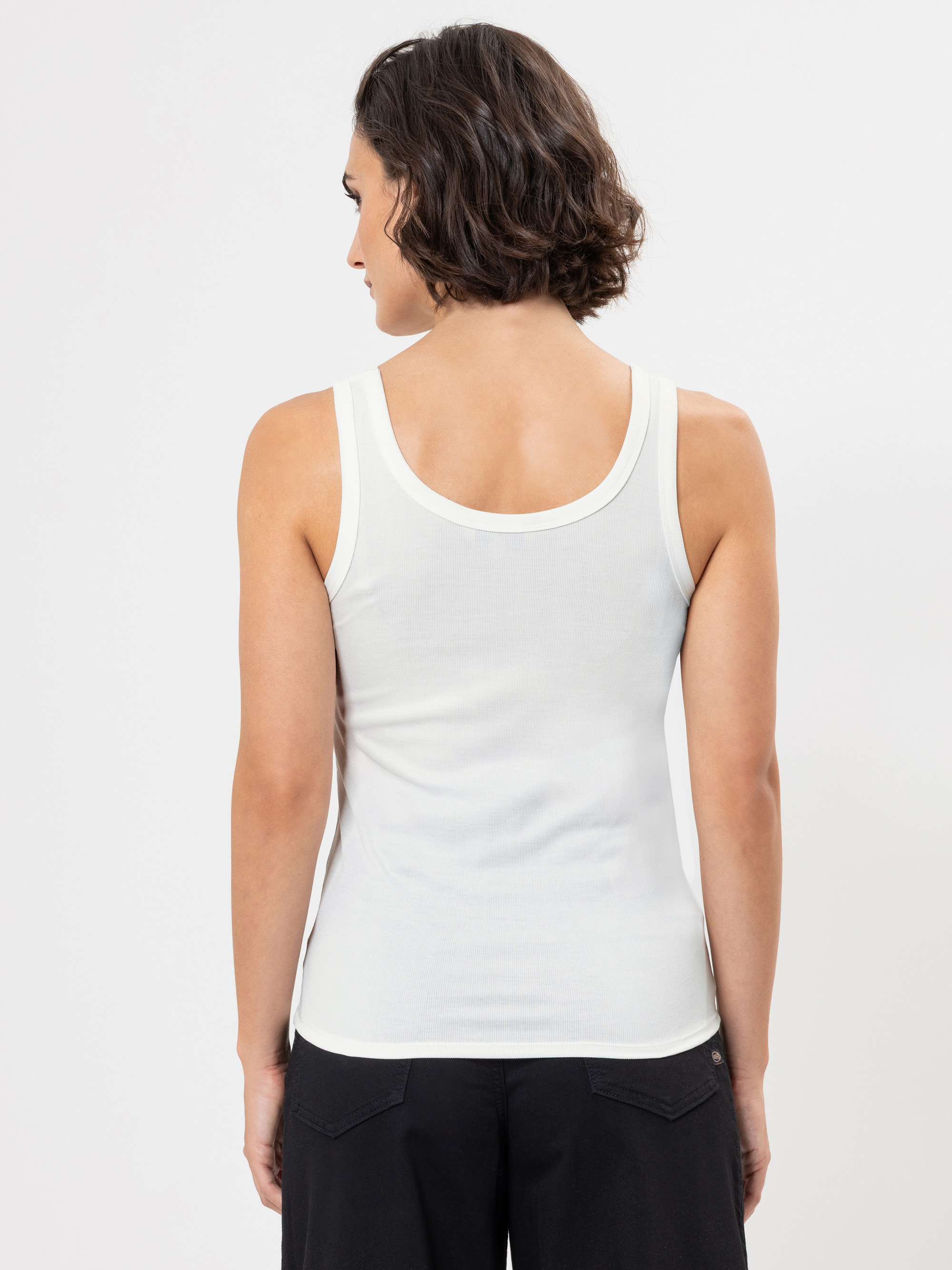 Eine Person mit kurzen, gewellten braunen Haaren wird von hinten gezeigt. Sie trägt ein weißes ärmelloses Tank-Top und eine schwarze Hose vor einem schlichten weißen Hintergrund.