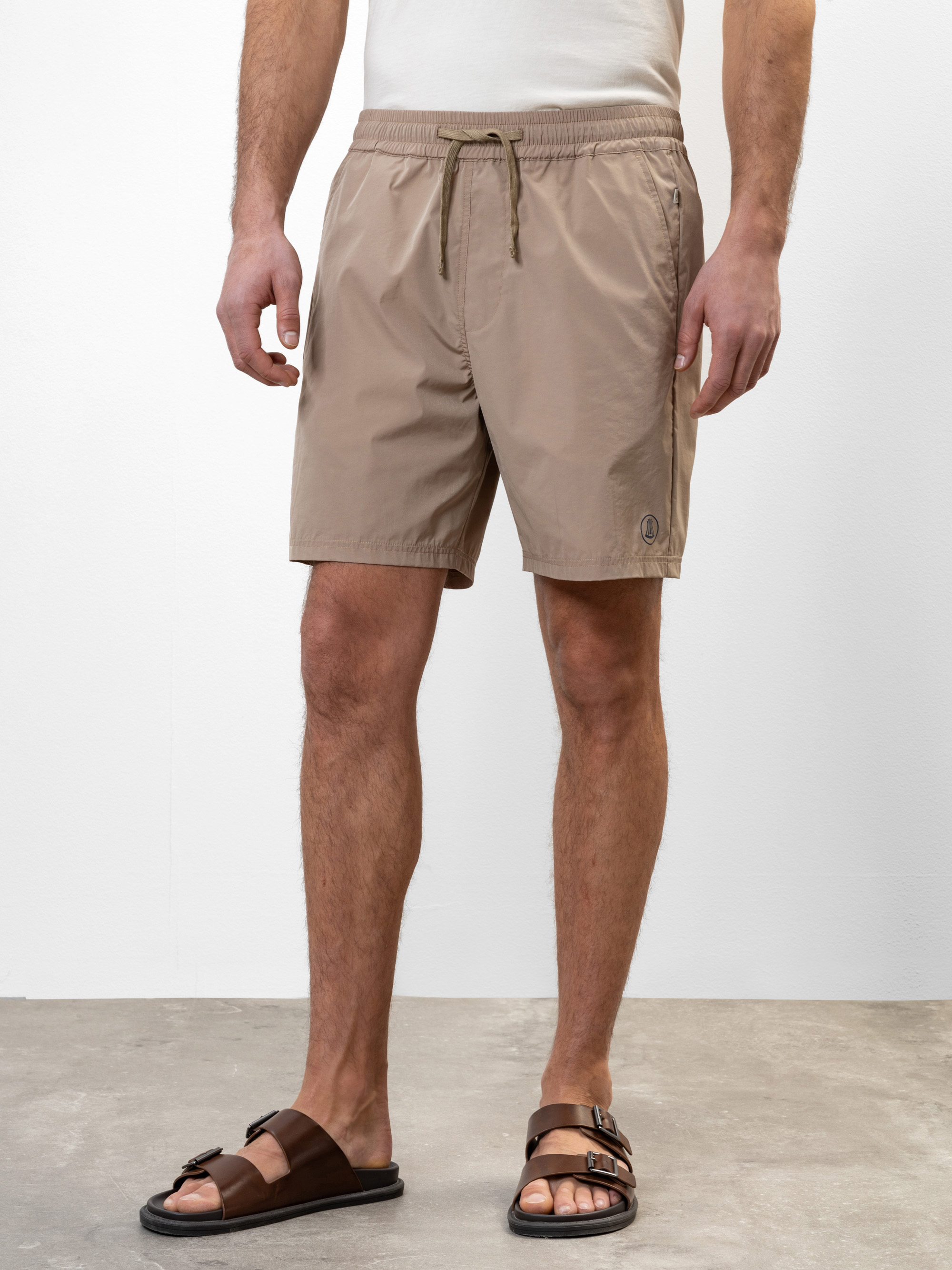 Eine Person, die ein weißes Hemd, beigefarbene Shorts und braune Sandalen trägt, steht auf einem grauen Boden vor einer schlichten weißen Wand. Das Foto ist so beschnitten, dass es von den Schultern bis zu den Füßen reicht.