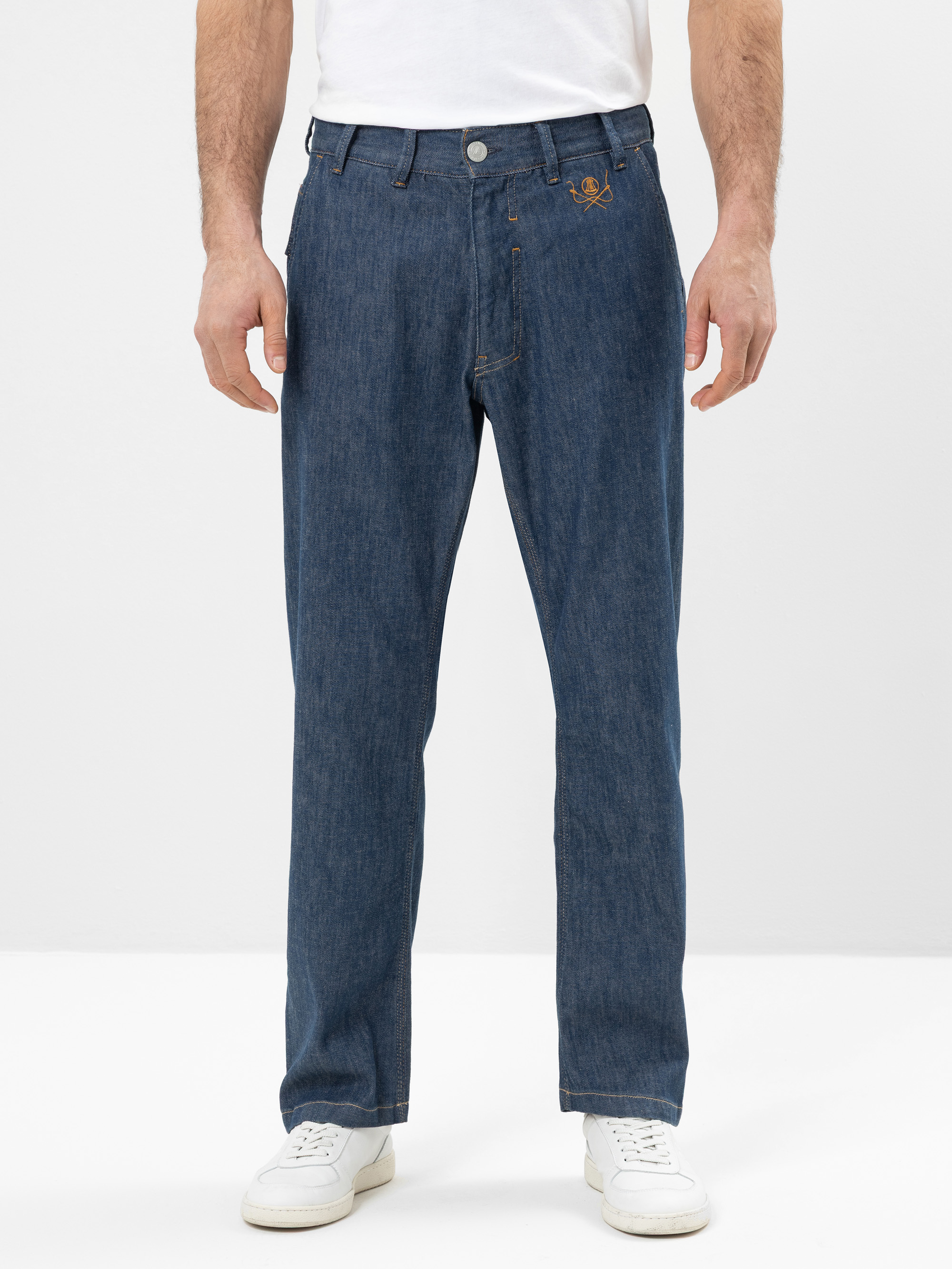 Ein Mann, der ein weißes T-Shirt, eine blaue Jeans mit geradem Bein und einem kleinen aufgestickten Logo in der Nähe der Hosentasche sowie weiße Turnschuhe trägt, steht vor einem schlichten weißen Hintergrund.