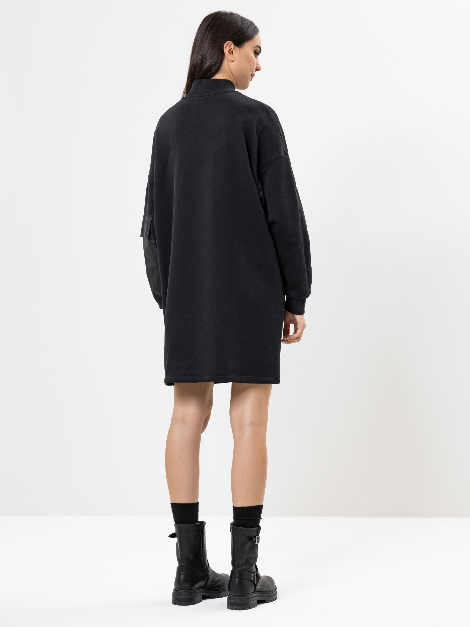 Eine Frau steht mit dem Gesicht zur Wand und trägt ein schwarzes, übergroßes Sweatshirtkleid und schwarze Kampfstiefel. Der Hintergrund ist einfarbig weiß, und ihr glattes dunkles Haar fällt ihr über die Schultern.