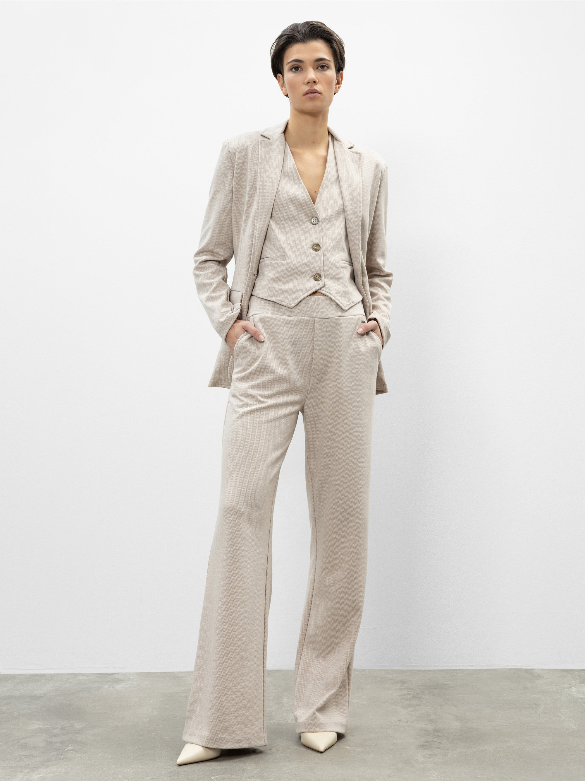 Eine Person steht vor einer schlichten weißen Wand und trägt einen hellbeigen dreiteiligen Anzug mit weit geschnittenen Hosen, einer zugeknöpften Weste und einem Blazer. Sie hat kurzes dunkles Haar und cremefarbene spitze Schuhe.