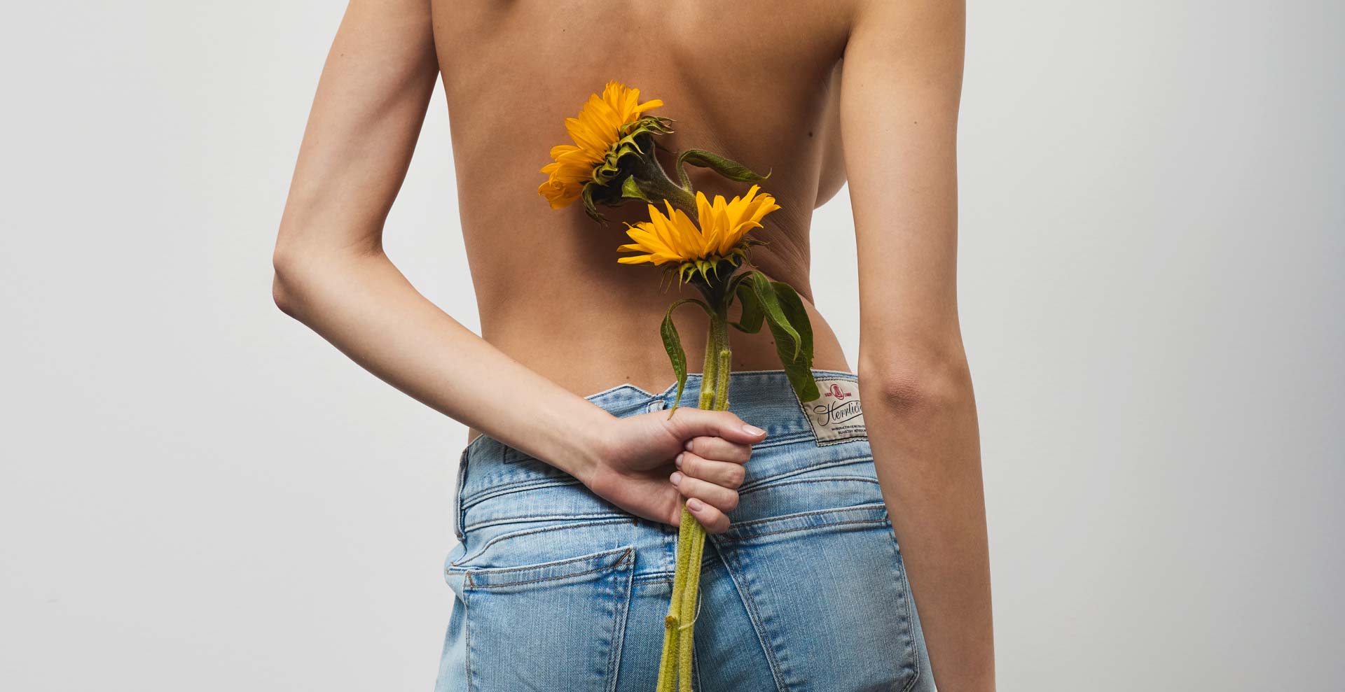 Eine Person mit nacktem Rücken, die eine blaue Jeans trägt, hält zwei gelbe Sonnenblumen hinter ihrem Rücken vor einem schlichten weißen Hintergrund.