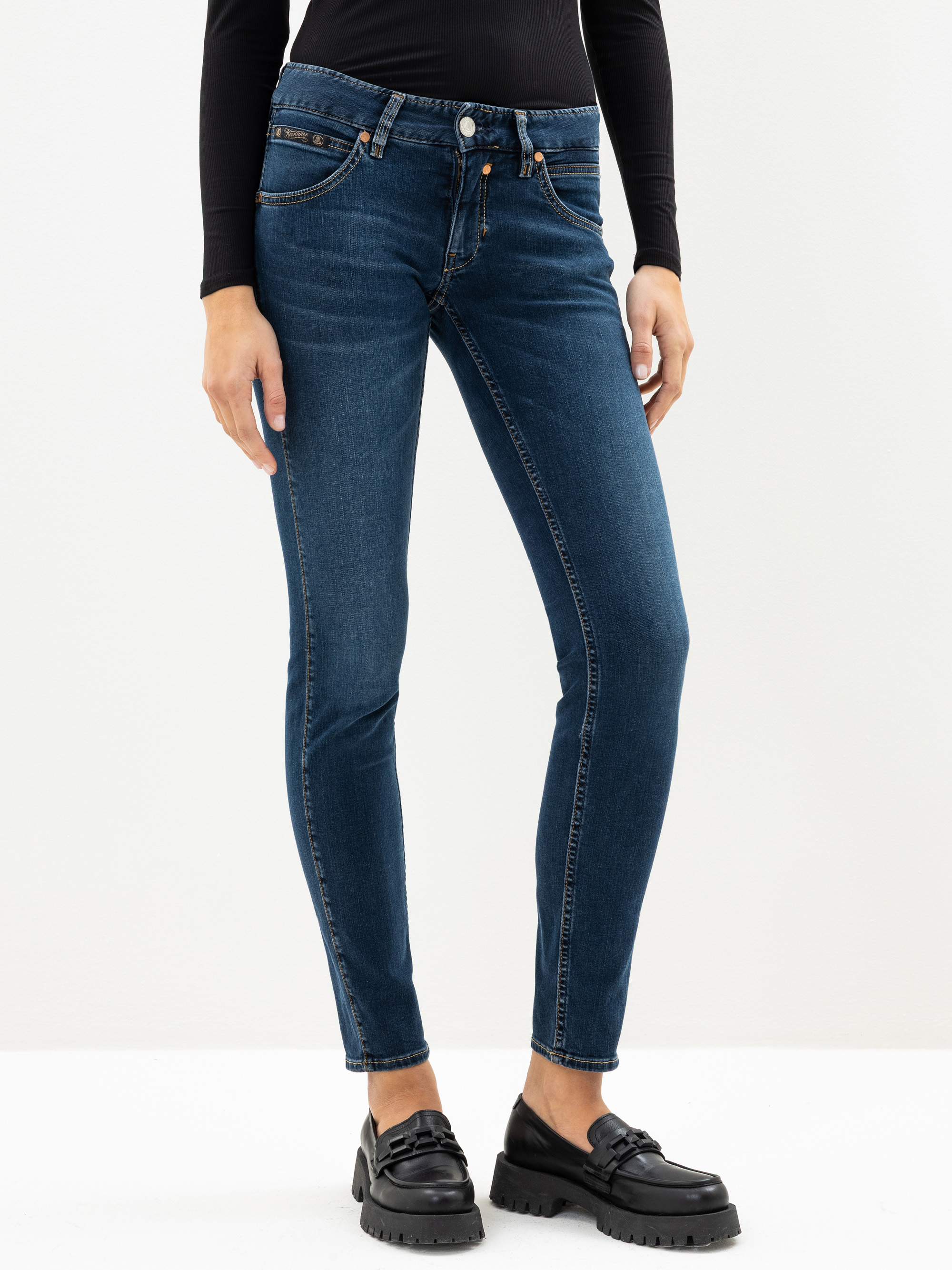 Person, die ein schwarzes langärmeliges Oberteil, blaue Röhrenjeans und schwarze Halbschuhe trägt und vor einem schlichten weißen Hintergrund steht. Nur der Körper von den Schultern bis zu den Füßen ist sichtbar.