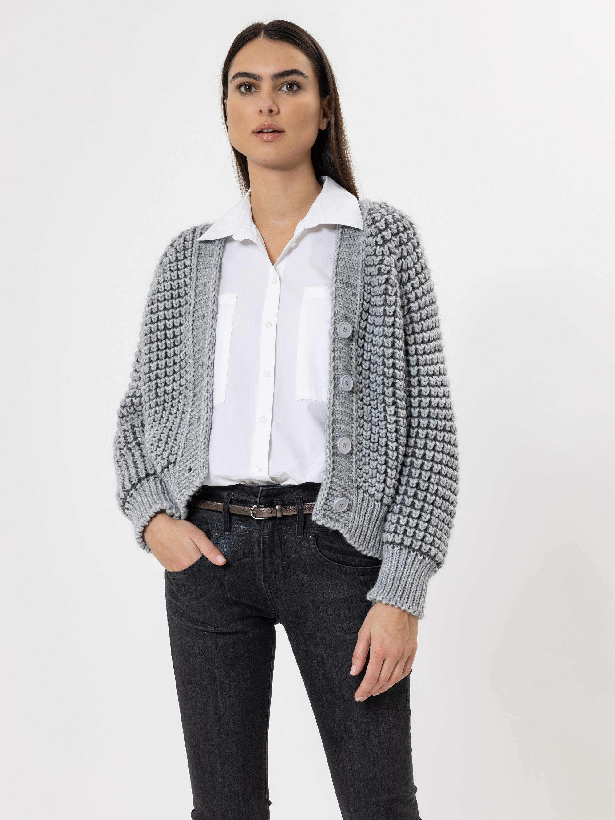 Eine Frau steht vor einem schlichten Hintergrund. Sie trägt eine dicke graue Strickjacke über einem weißen Button-up-Hemd, dazu eine schwarze Jeans. Eine Hand steckt in ihrer Tasche und sie blickt leicht zur Seite.
