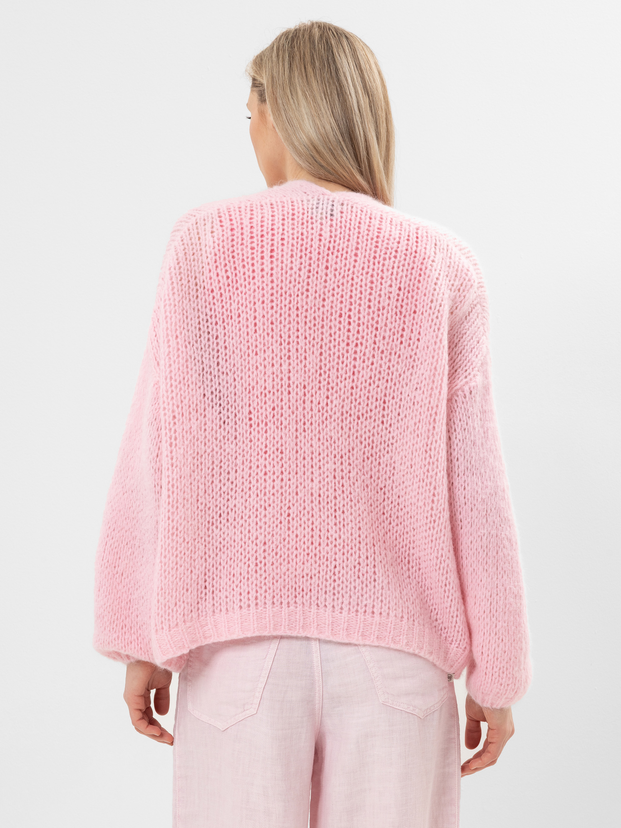 Eine Person mit langem, hellbraunem Haar, die einen dicken, hellrosa Strickpullover und eine dazu passende hellrosa Hose trägt, dargestellt von hinten vor einem schlichten, weißen Hintergrund.