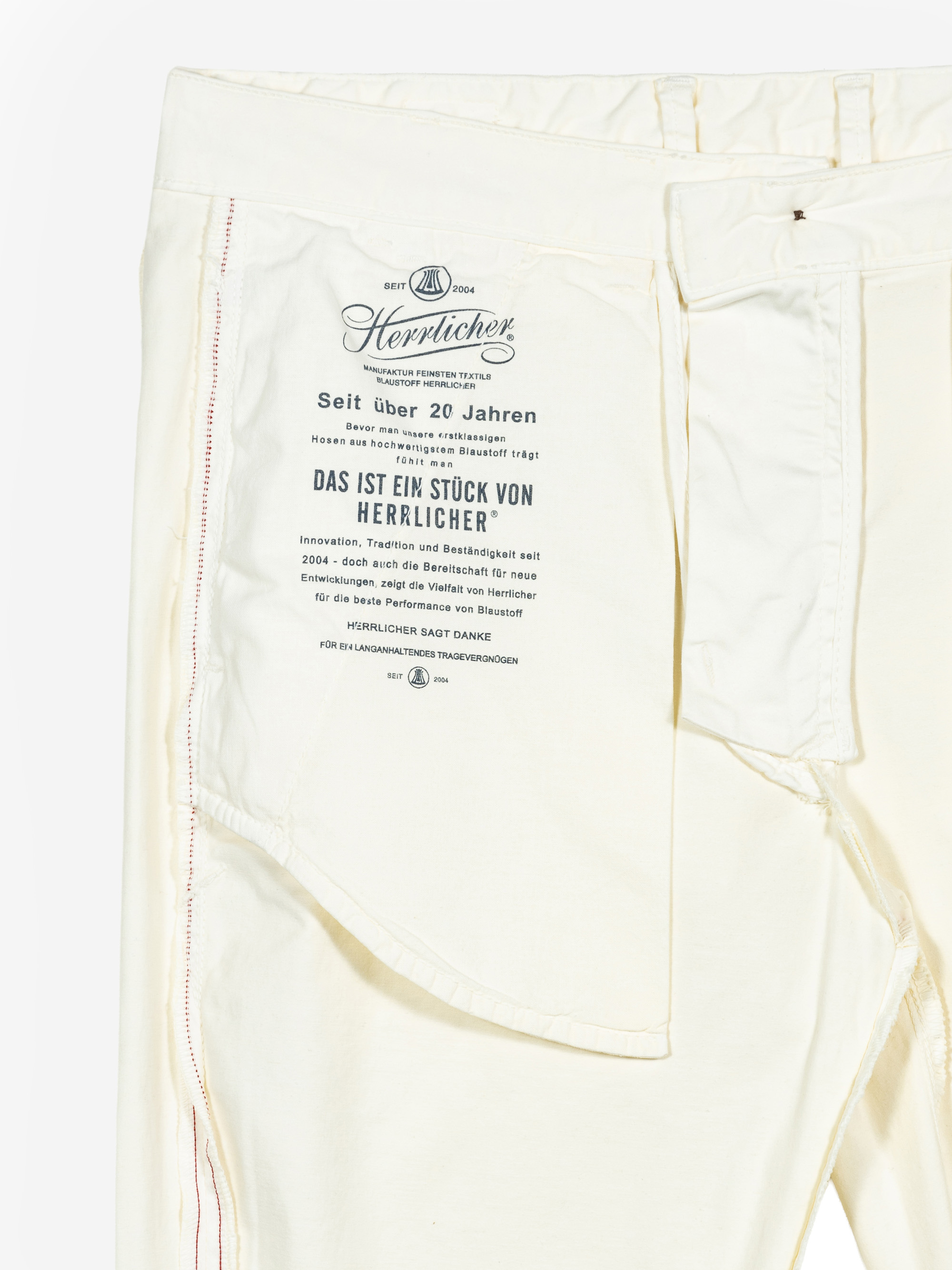Off-White-Hose mit umgedrehter Innenseite, die ein großes, auf Deutsch gedrucktes Etikett auf dem Innenstoff zeigt, das die Marke Herrlicher und ihre Geschichte beschreibt, sowie sichtbare Nähte und eine Seitentasche.