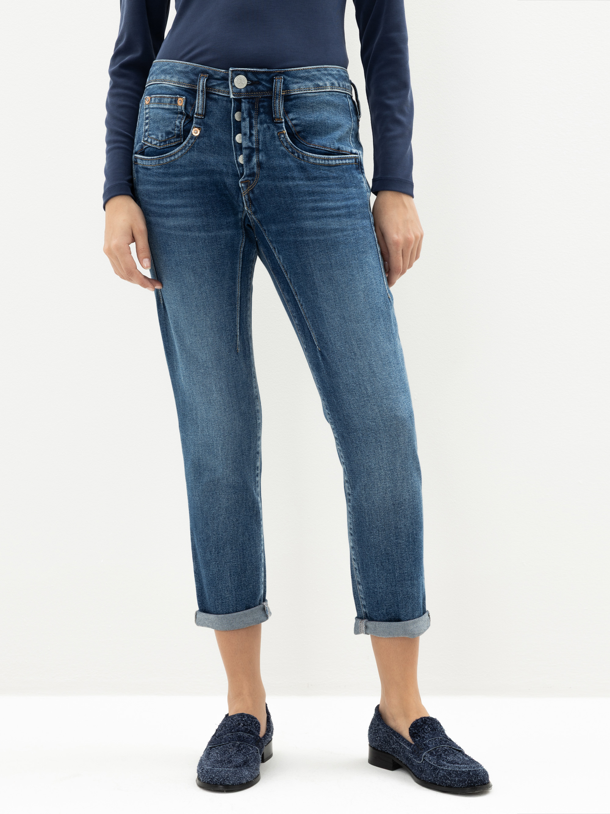Person, die eine blaue Jeans mit Manschetten, ein langärmeliges marineblaues Oberteil und marineblaue Halbschuhe trägt und vor einem schlichten hellen Hintergrund steht. Nur der Unterkörper ist sichtbar.