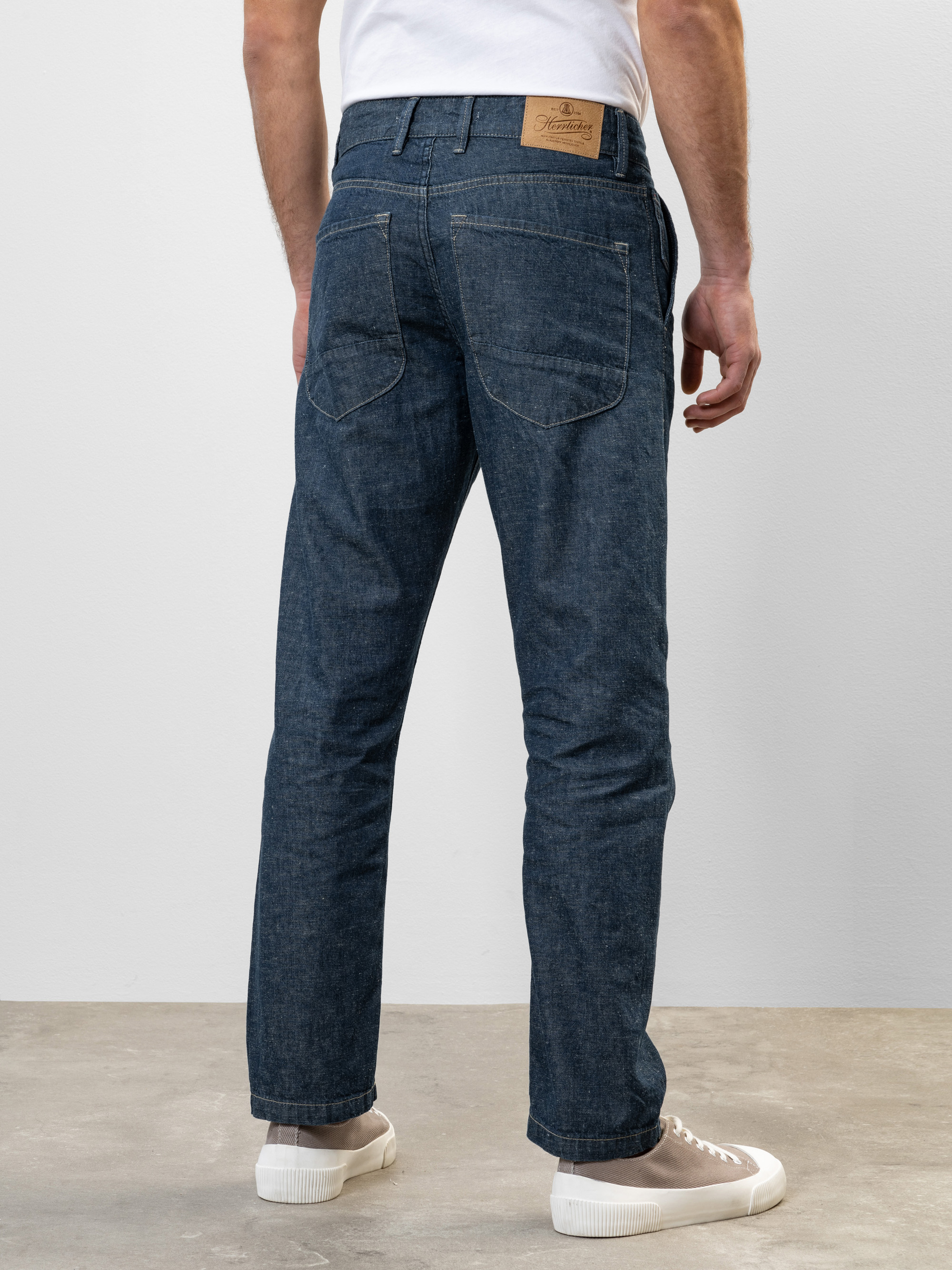 Ein Mann, der ein weißes T-Shirt, eine blaue Jeans und weiße Turnschuhe trägt, steht mit dem Gesicht zur Seite und zeigt die Rückseite der Jeans mit sichtbaren Gesäßtaschen und einem braunen Lederaufnäher am Bund.
