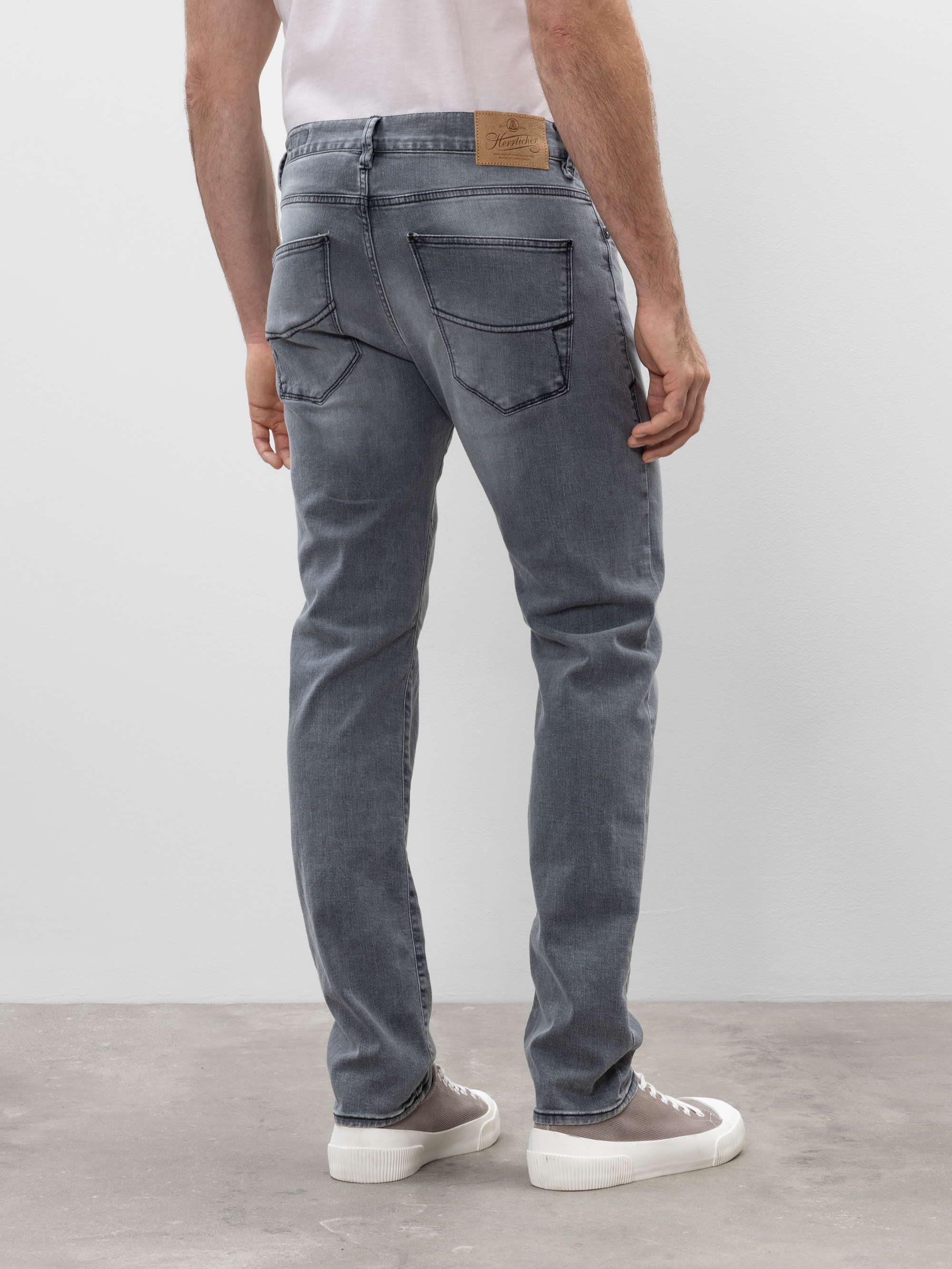 Ein Mann in hellgrauen Jeans und weißen Turnschuhen steht auf einem Betonboden, den Blick von der Kamera abgewandt, die Arme entspannt an den Seiten.