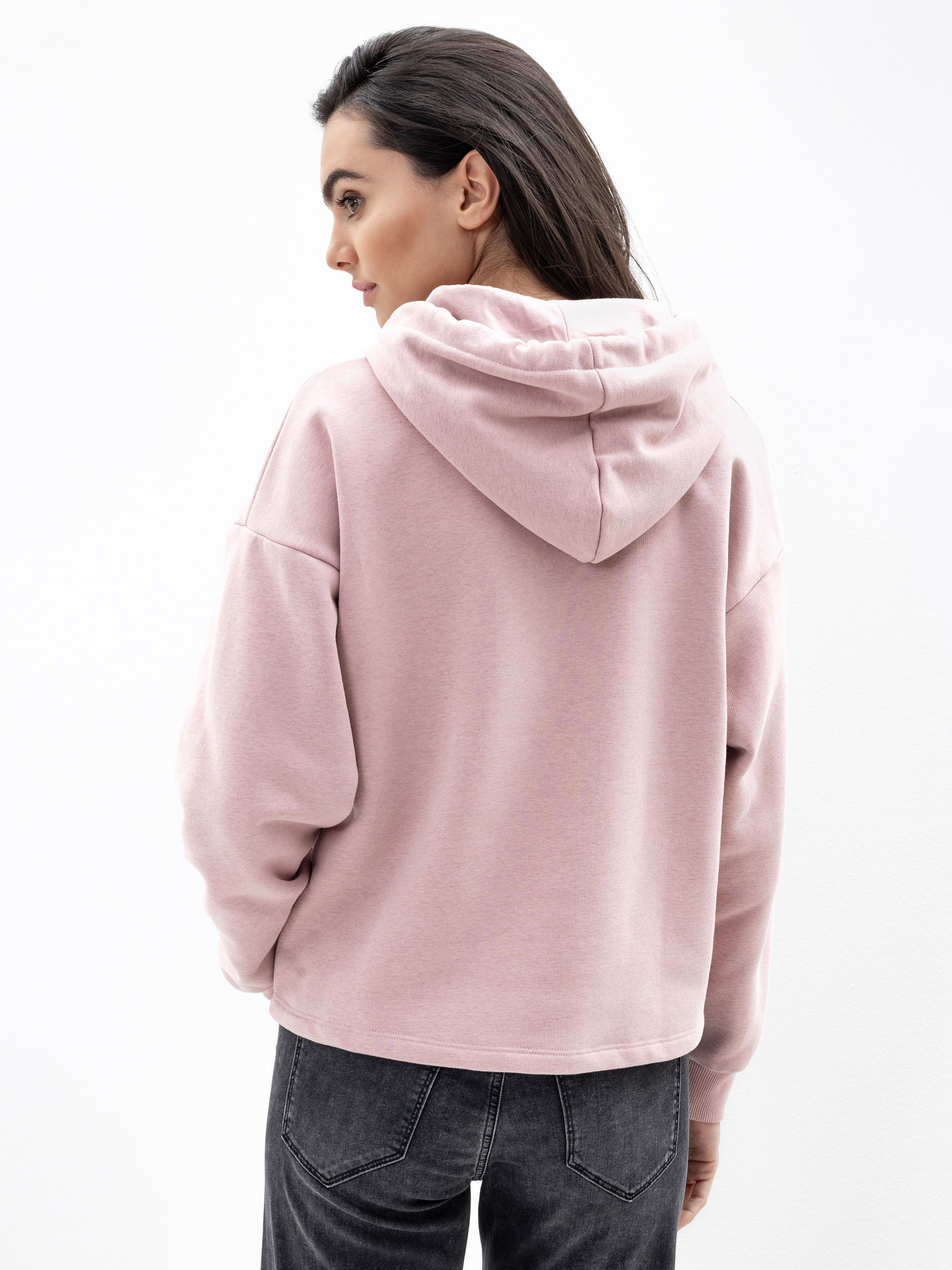 Eine Frau mit langen dunklen Haaren trägt ein hellrosa Kapuzensweatshirt und dunkle Jeans. Sie steht vor einem schlichten weißen Hintergrund und blickt mit leicht zur Seite gedrehtem Kopf zur Seite.