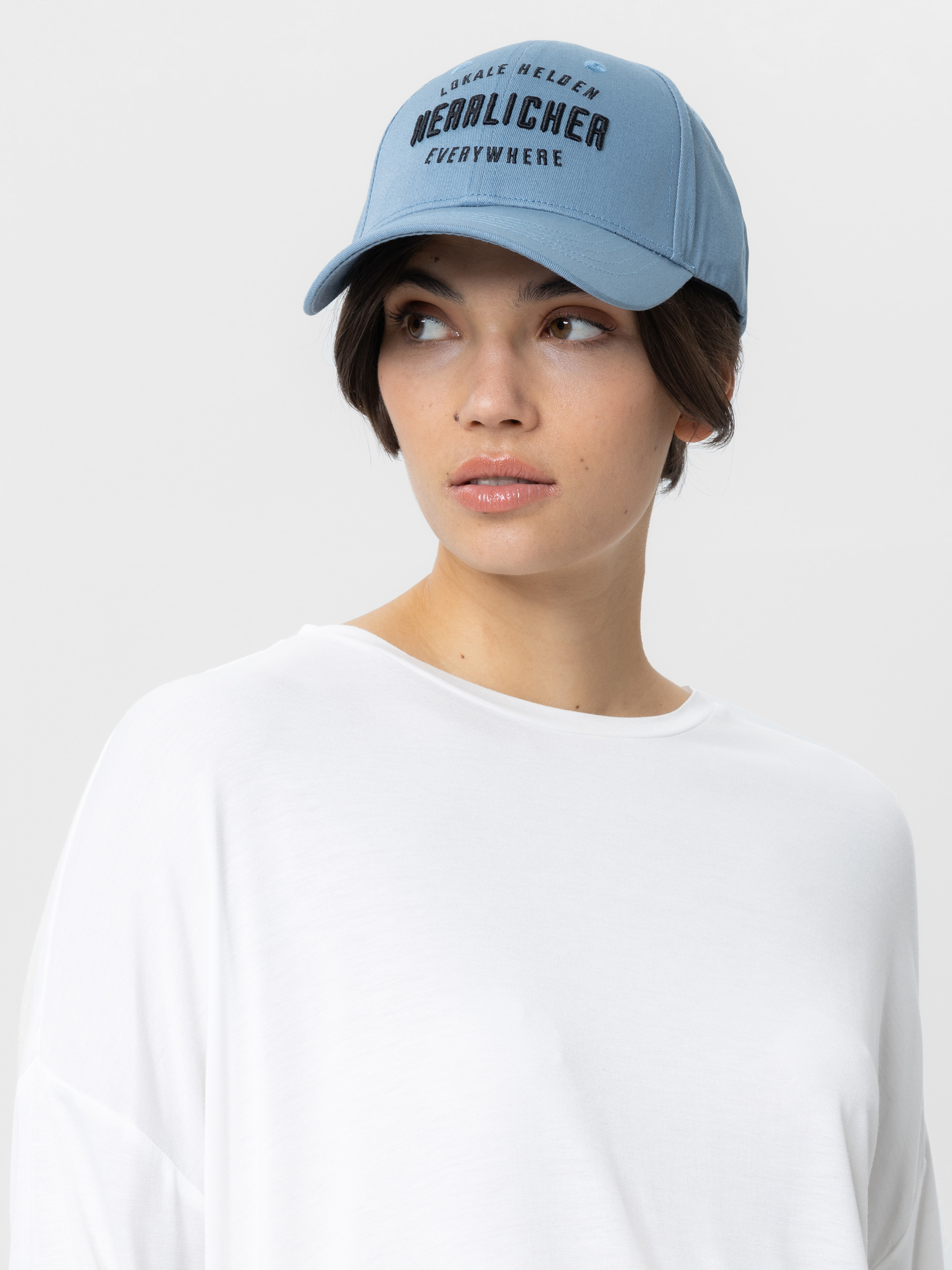 Eine Frau, die eine hellblaue Baseballkappe mit dunkler Aufschrift und ein lockeres weißes Oberteil trägt, schaut vor einem schlichten, hellen Hintergrund leicht nach rechts.